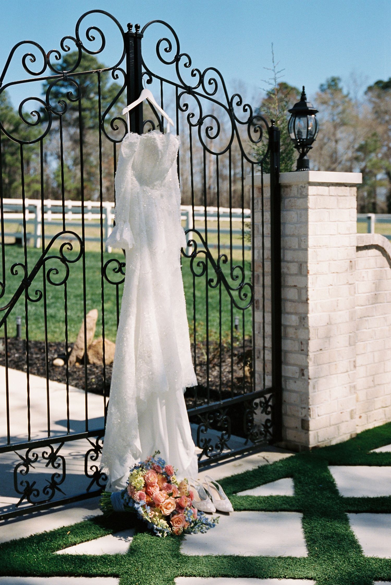 White wedding dress hanging on ornate black iron gate with brick pillars and green grass in an outdoor garden setting.