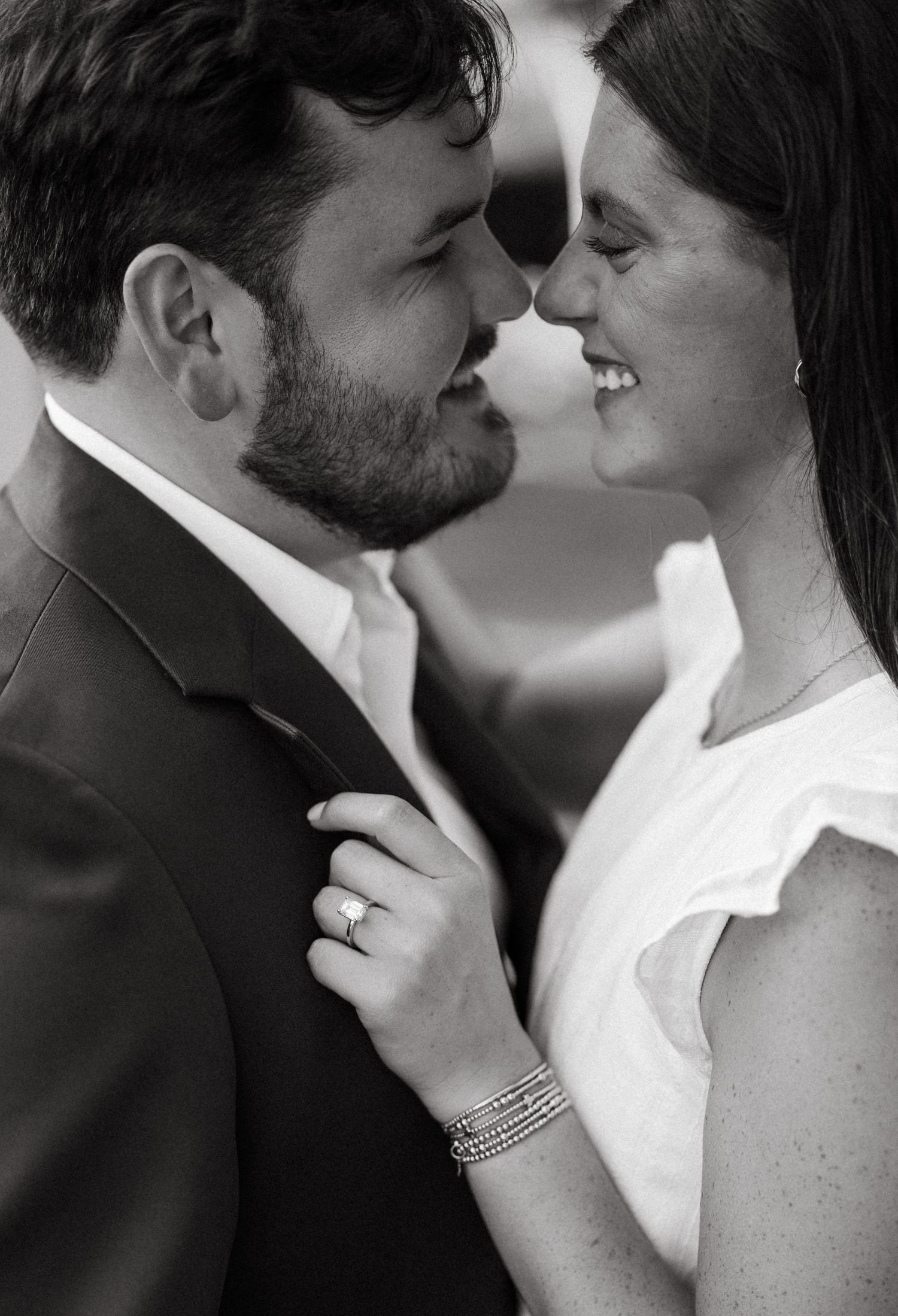 Black and white photo of a smiling couple in formal wear touching foreheads, woman wearing an engagement ring and bracelet.