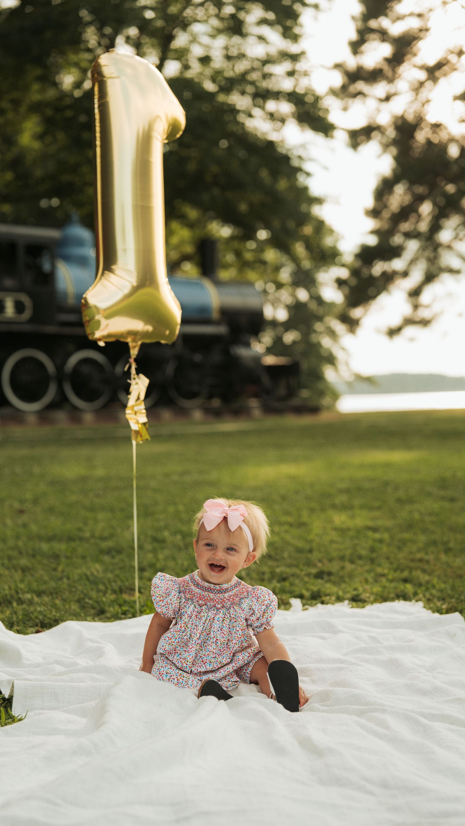 Baby in floral dress sitting on white blanket with gold number one balloon.
