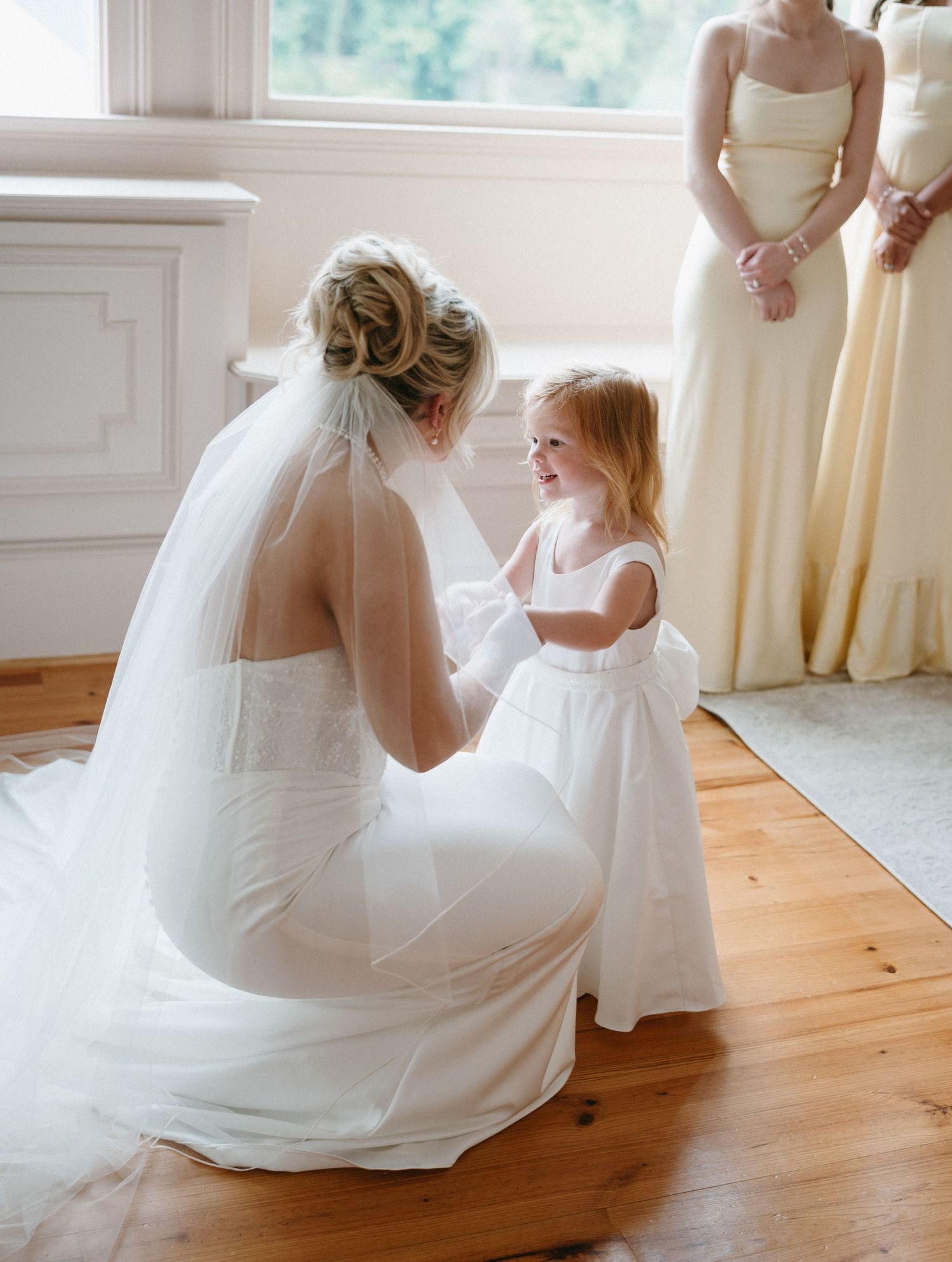 Bride in white gown kneels to adjust flower girl's dress while bridesmaids in yellow stand nearby on hardwood floor.