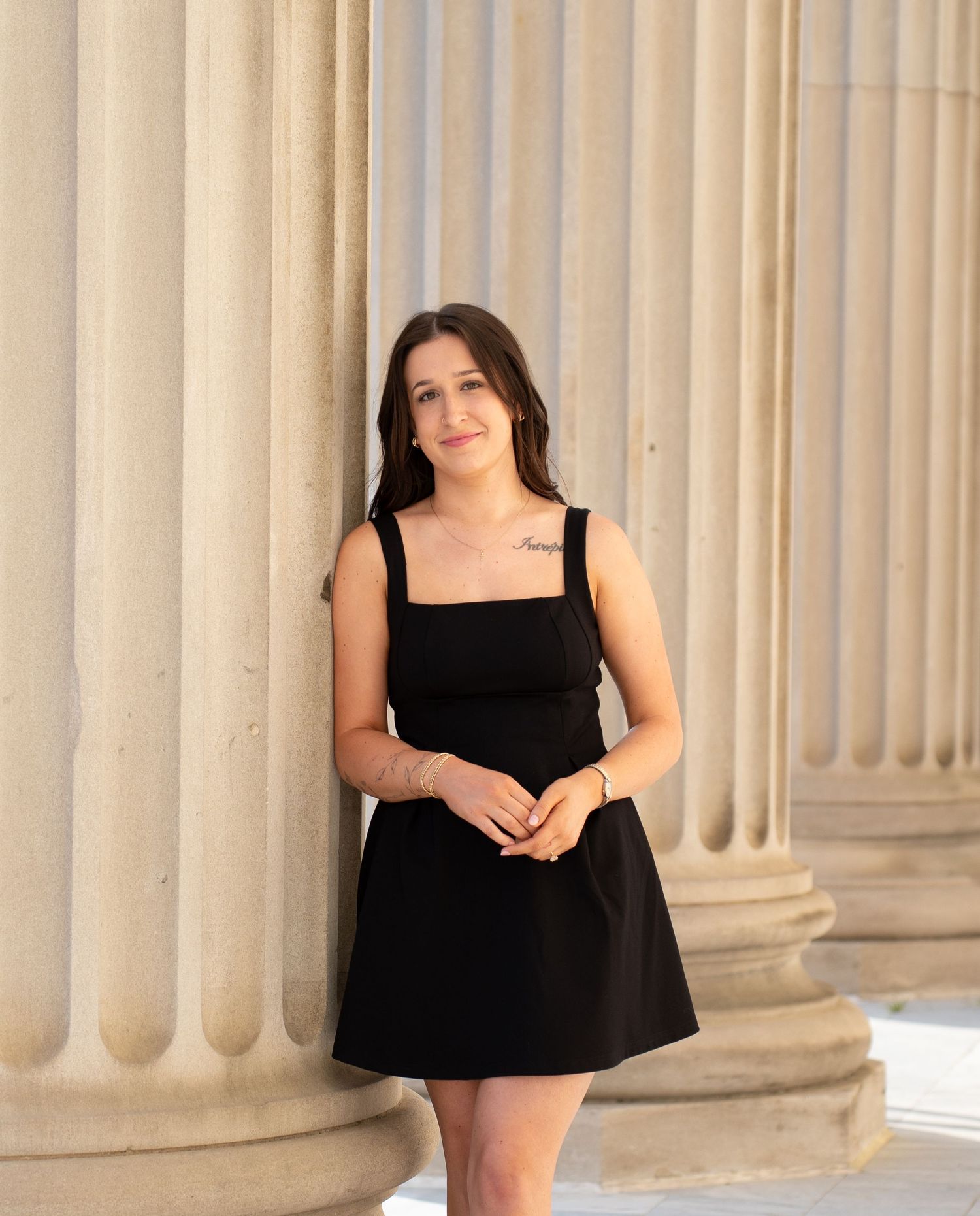 Young woman in black mini dress posing by stone columns outdoors, featuring a small tattoo on her chest.