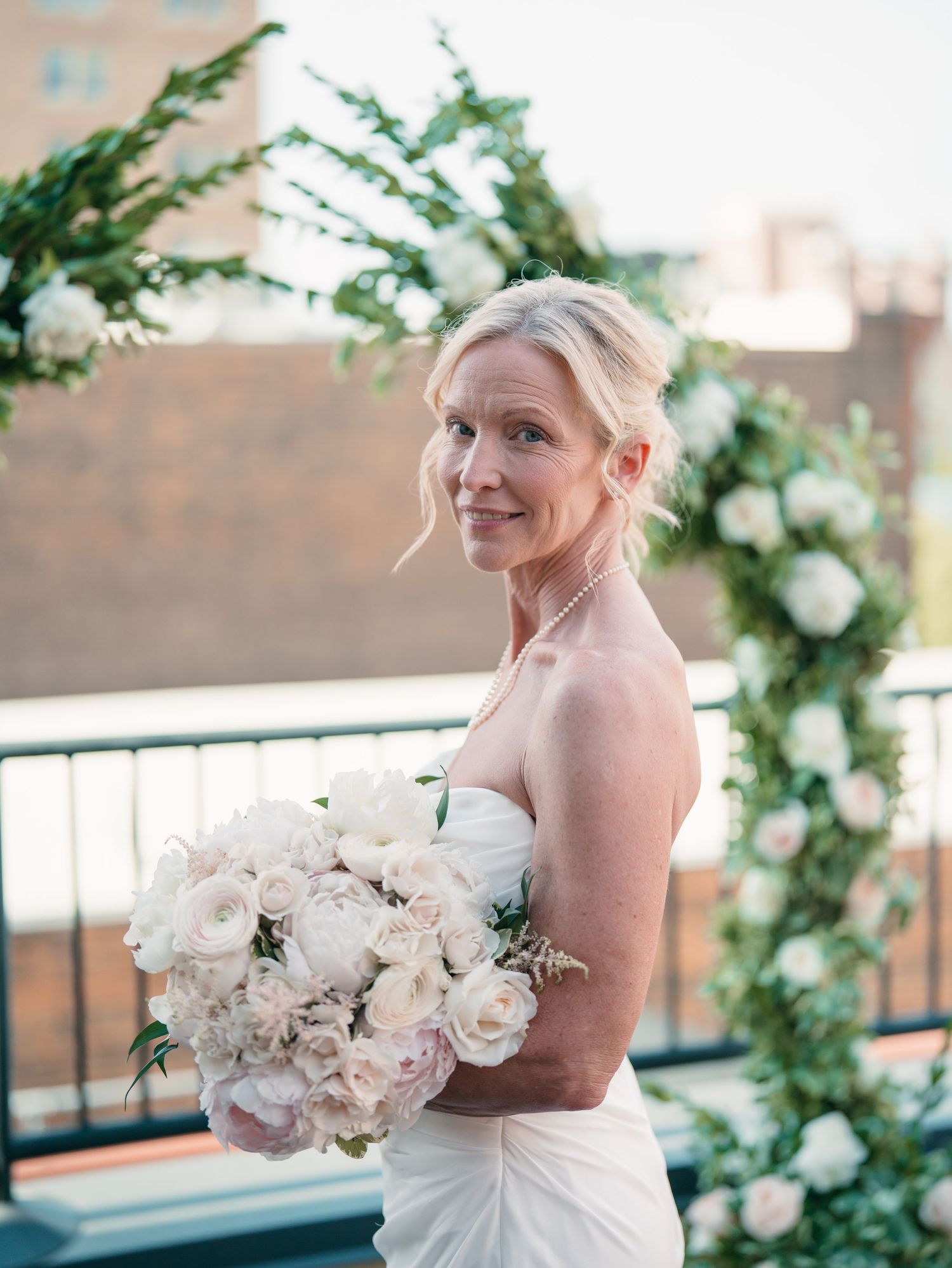 Bride in strapless white gown holding lush white rose bouquet, smiling on rooftop with floral arch backdrop.