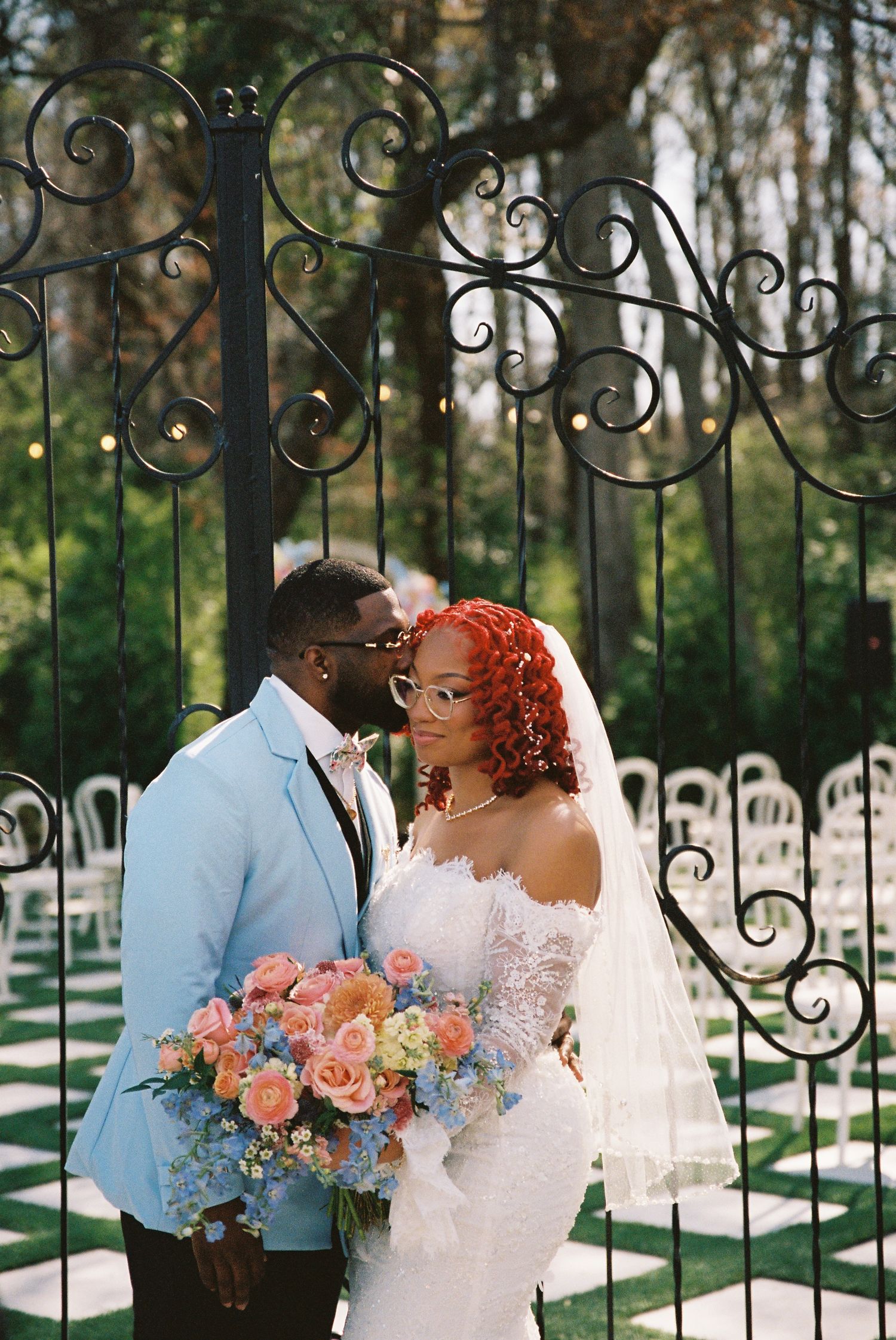 A romantic wedding couple shares intimate moments by an ornate iron gate with a pastel floral bouquet.