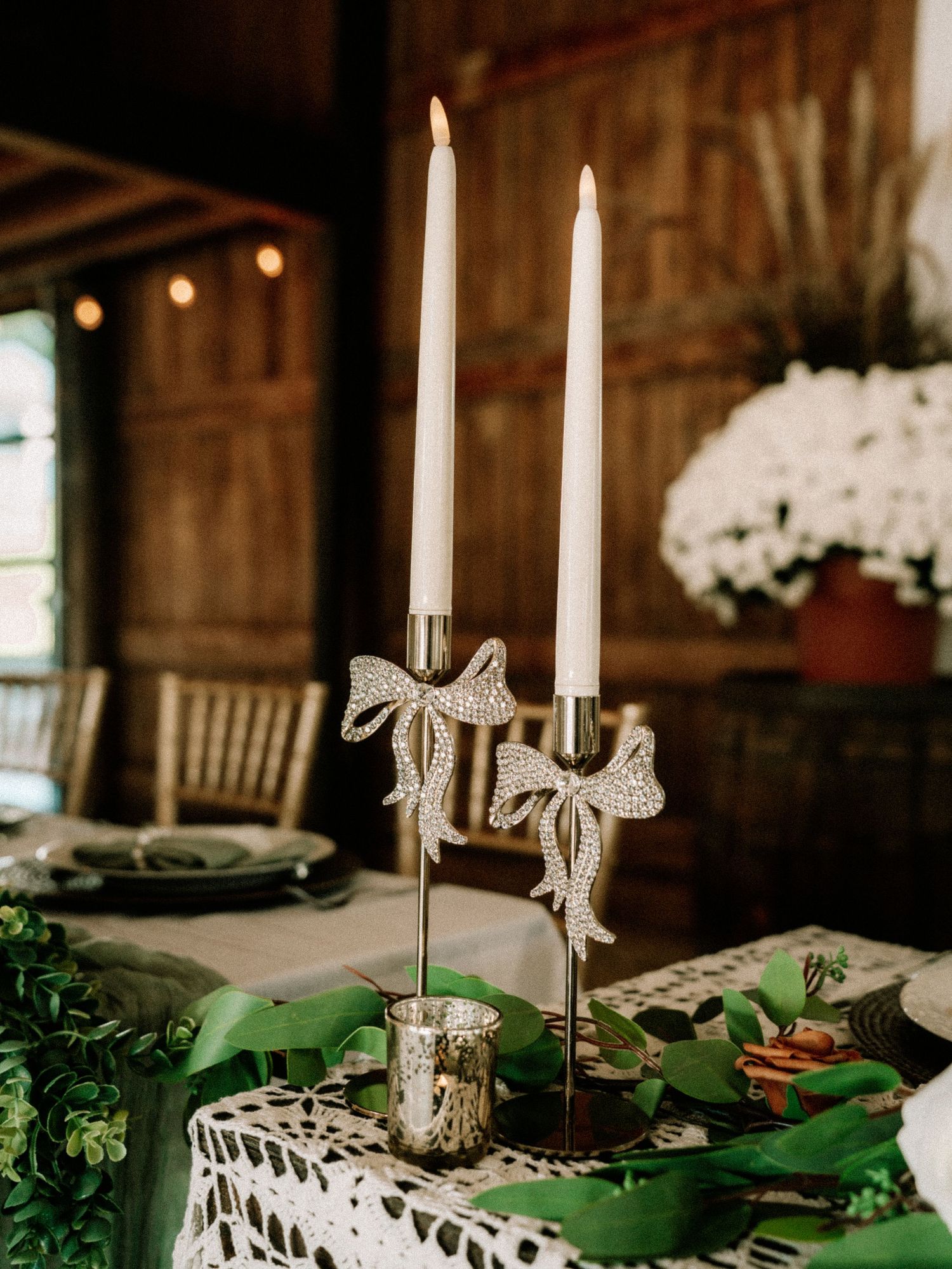 White taper candles with delicate bow decorations line an elegant rustic wedding table setting with greenery accents.