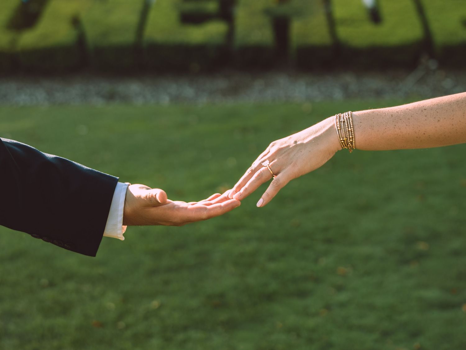Two people reaching toward each other, one wearing a suit and the other a gold bracelet and ring, with green grass behind.