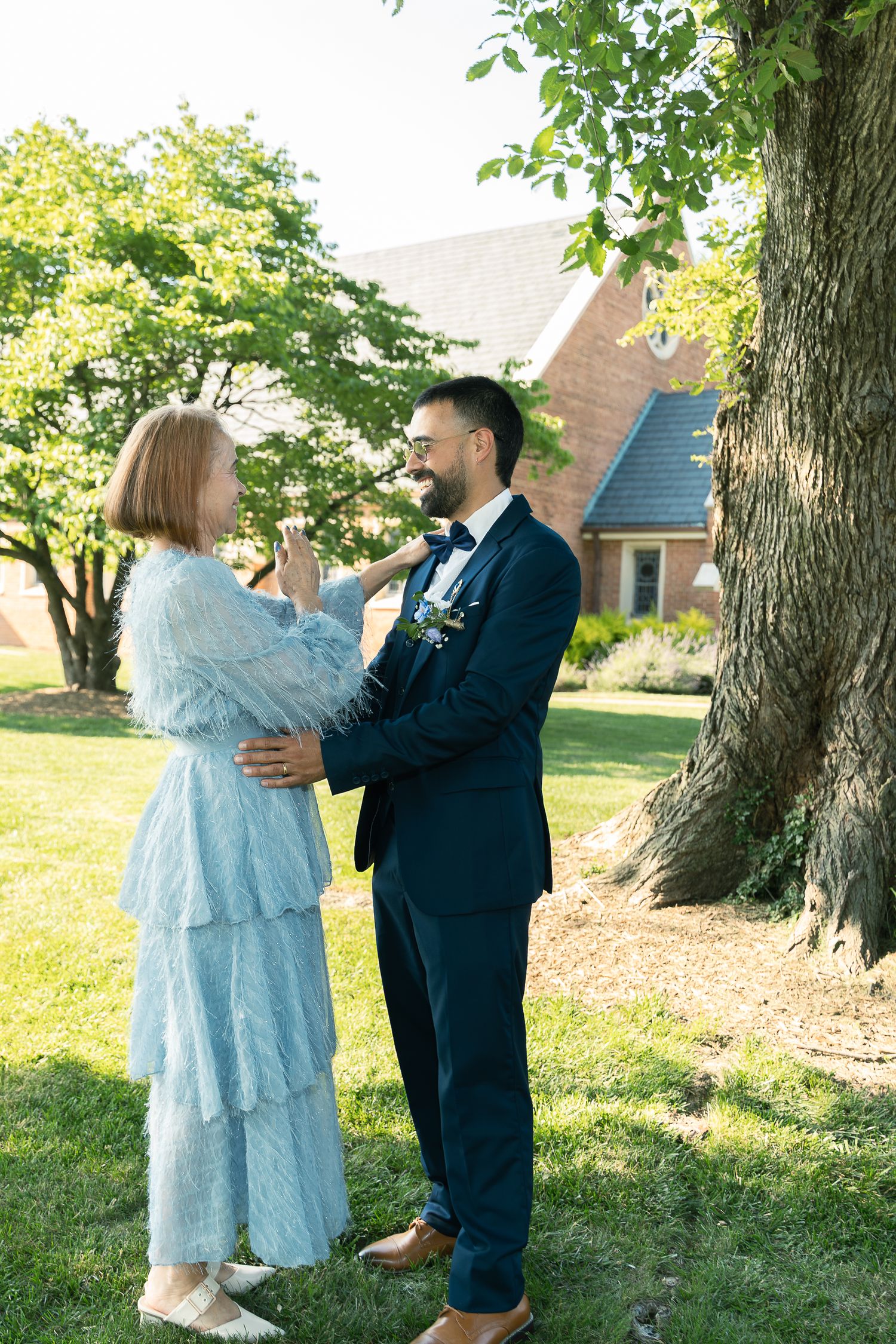 A couple in formal attire share an intimate moment under a large tree on a sunny day near a brick building.