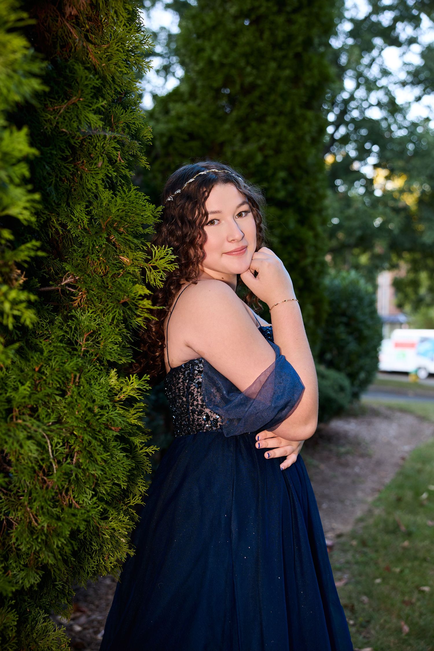 A person in a navy blue formal dress poses thoughtfully against a backdrop of evergreen trees in an outdoor setting.