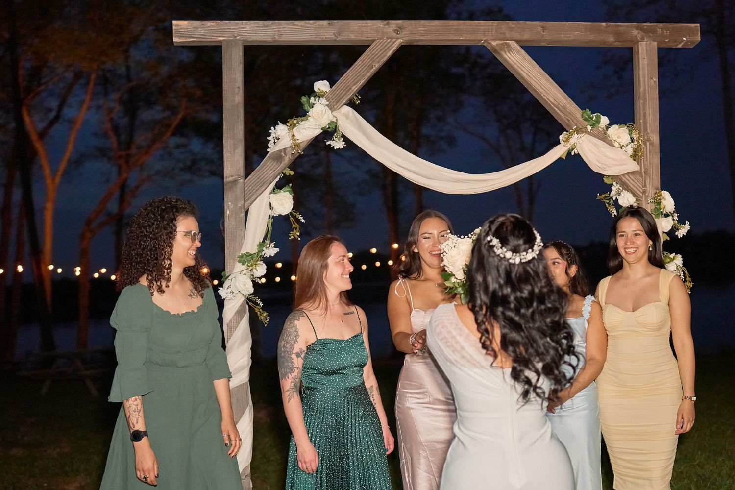 Bride in white gown laughing with bridesmaids under rustic wooden arch adorned with white flowers at outdoor evening wedding.
