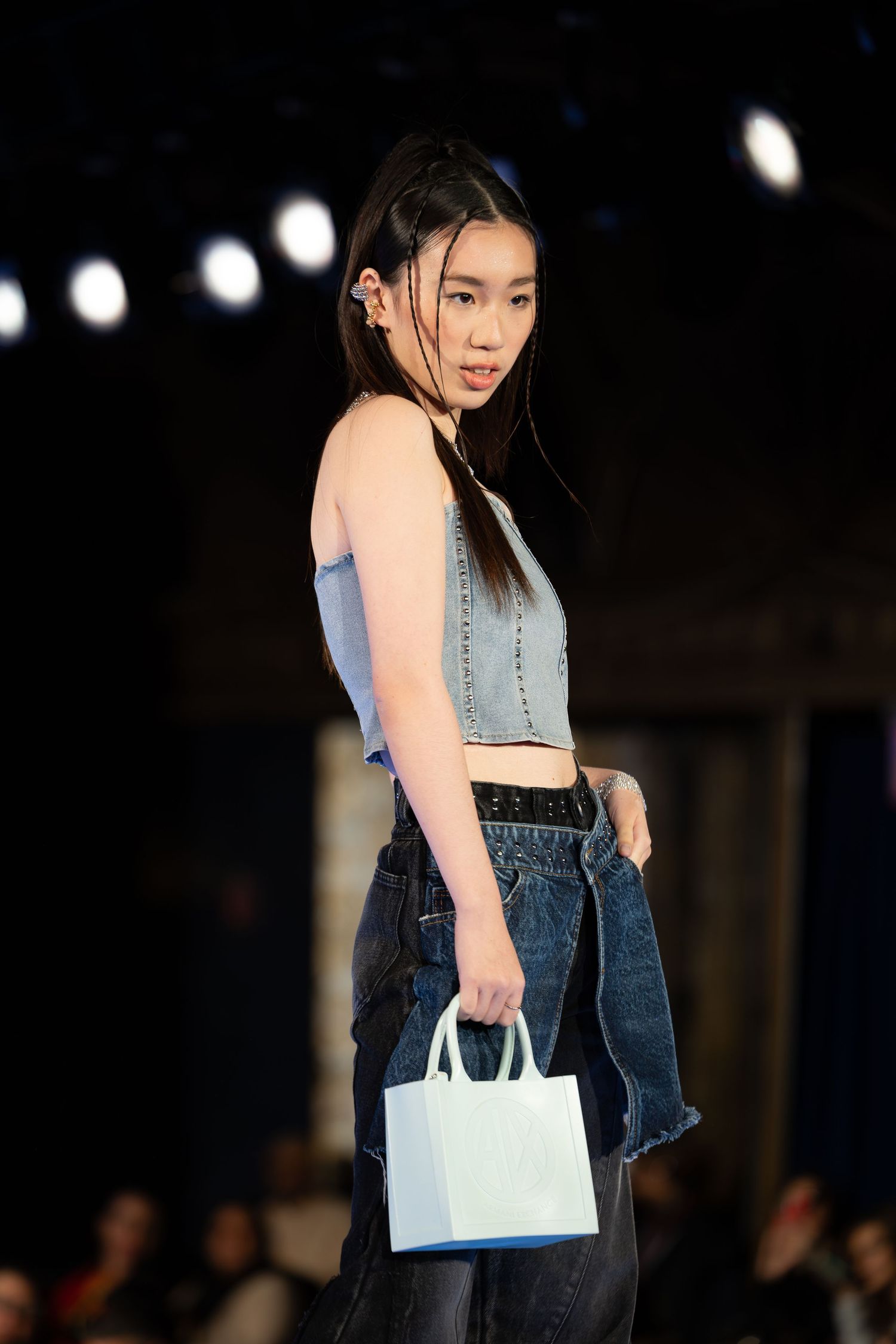 Fashion model walks runway in cropped blue top and jeans while carrying small white handbag under dramatic lighting.