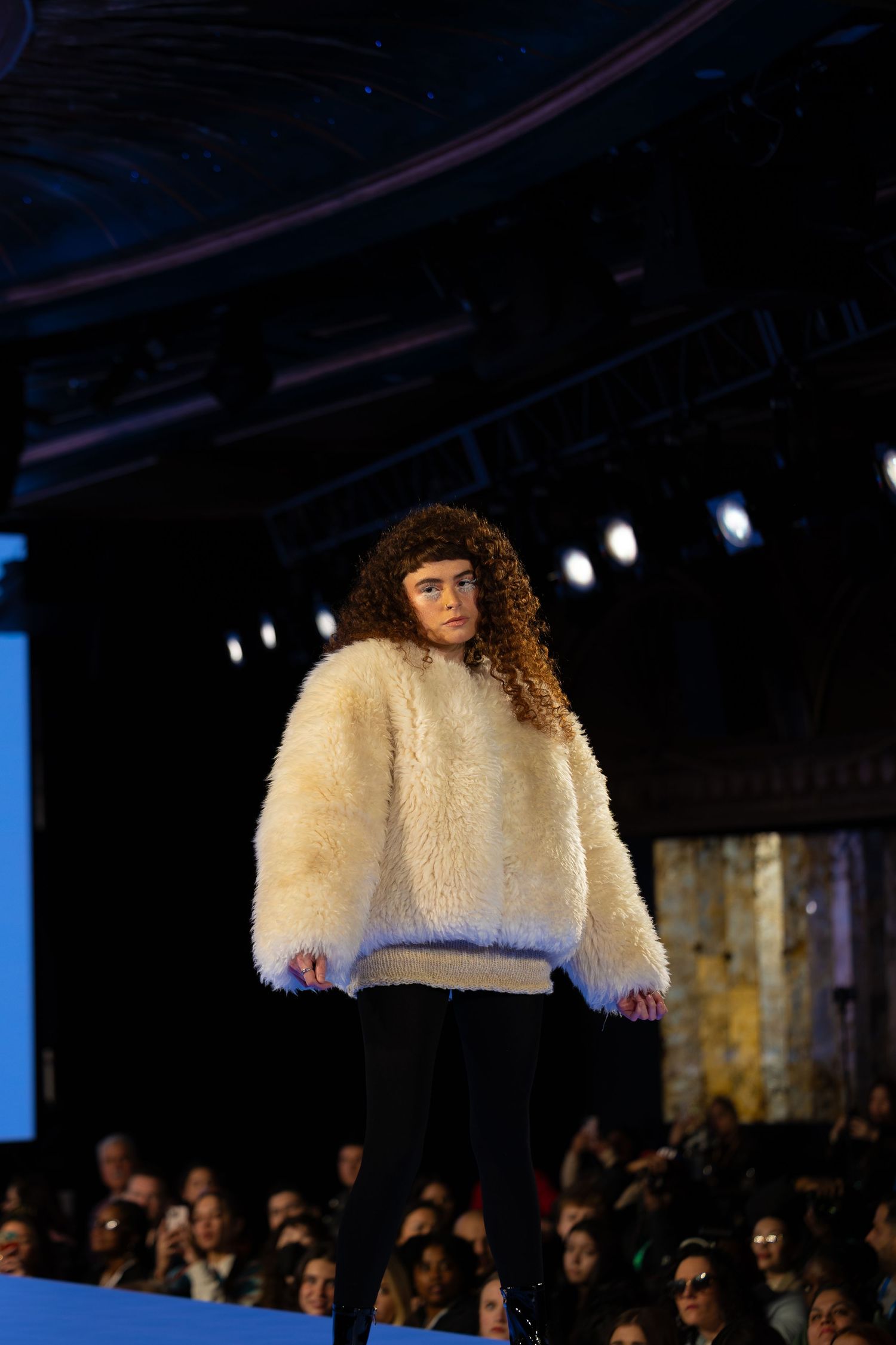 Model wearing fluffy cream colored sweater and black pants on illuminated runway.