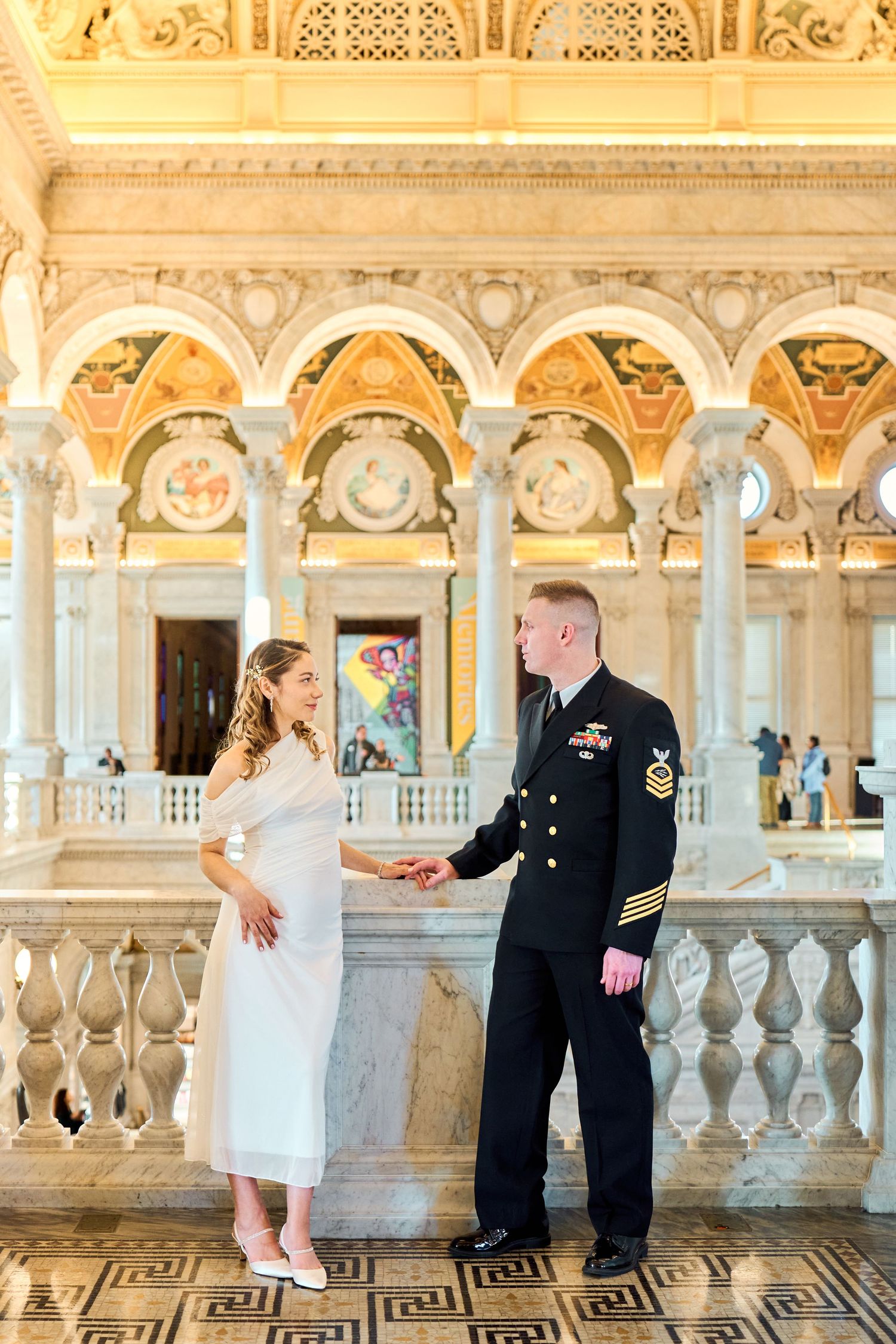 Pregnant woman in white dress holds hands with Navy officer in ornate marble library hall.