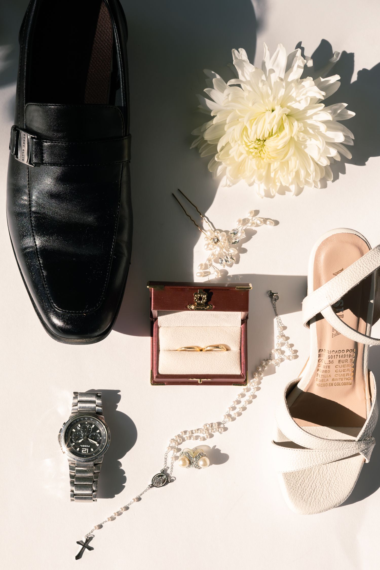Wedding accessories including black dress shoe, pearl necklace, white flower, engagement ring box, wristwatch and bridal heel.