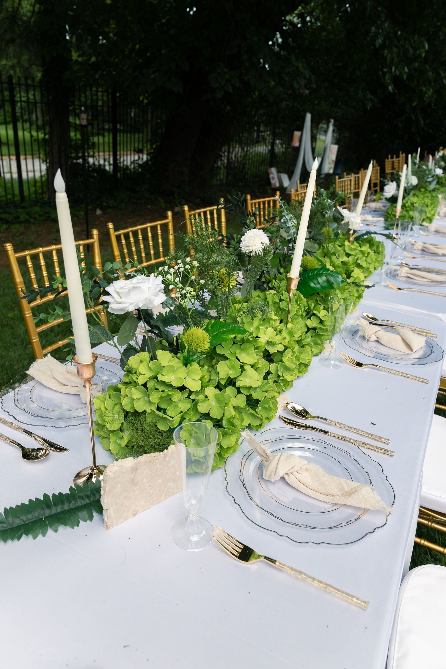 Elegant table settings with white linens, gold chairs, and fresh greenery featuring clear glass plates and white candles.