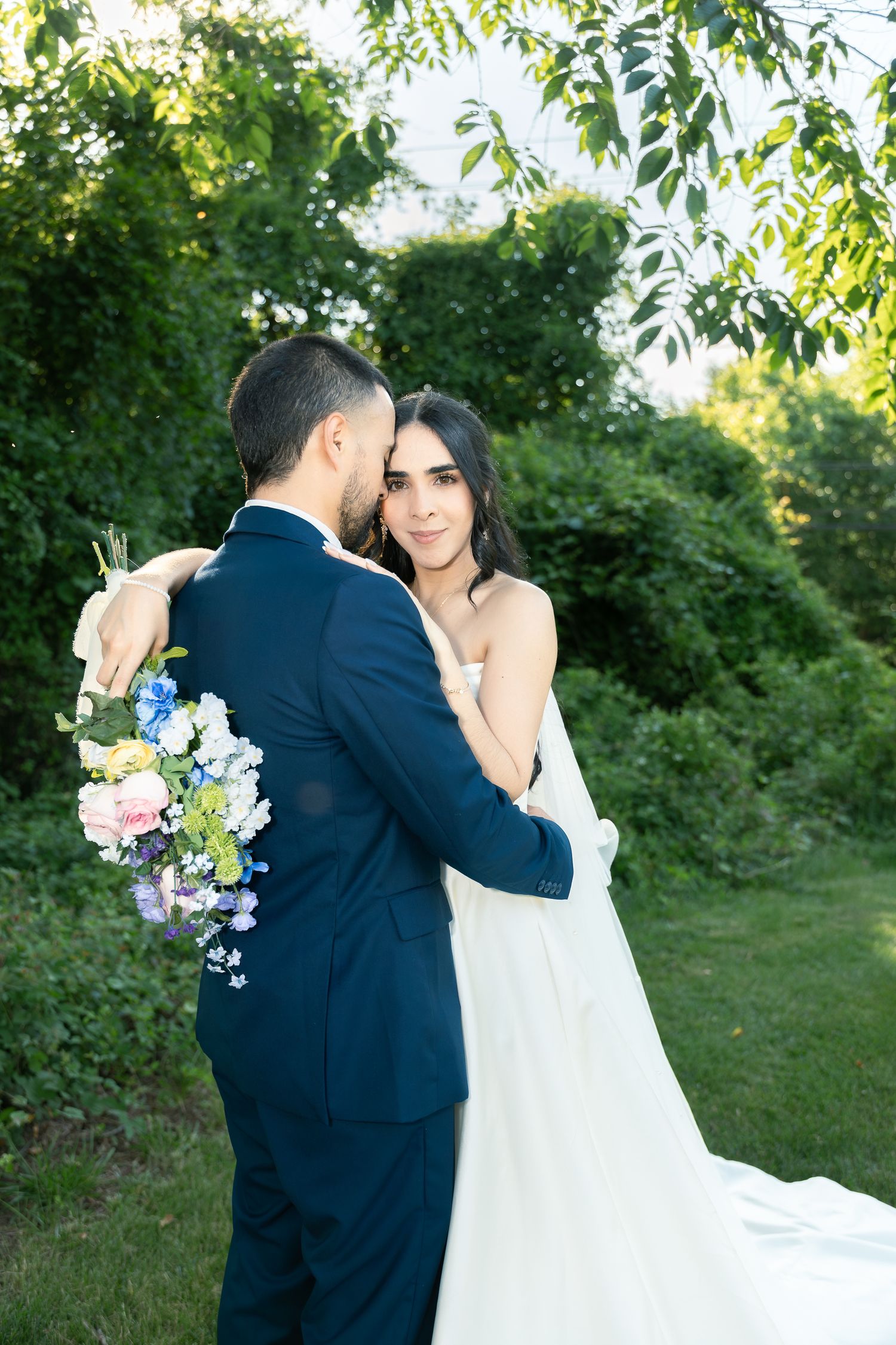 Romantic wedding photo of couple embracing outdoors with bouquet of blue and white flowers in garden setting.