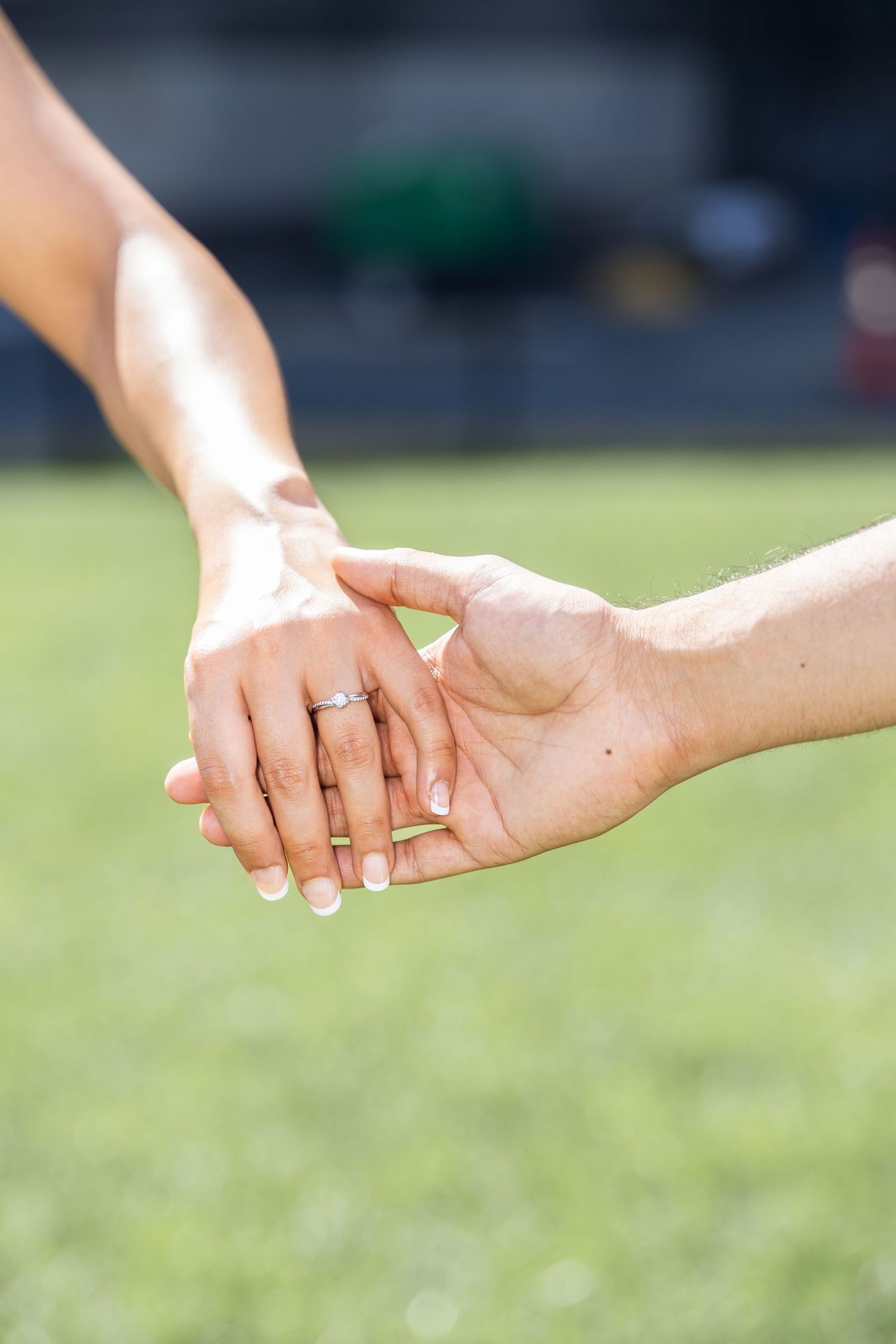 Two hands gently touch while displaying an engagement ring against a blurred green outdoor background.