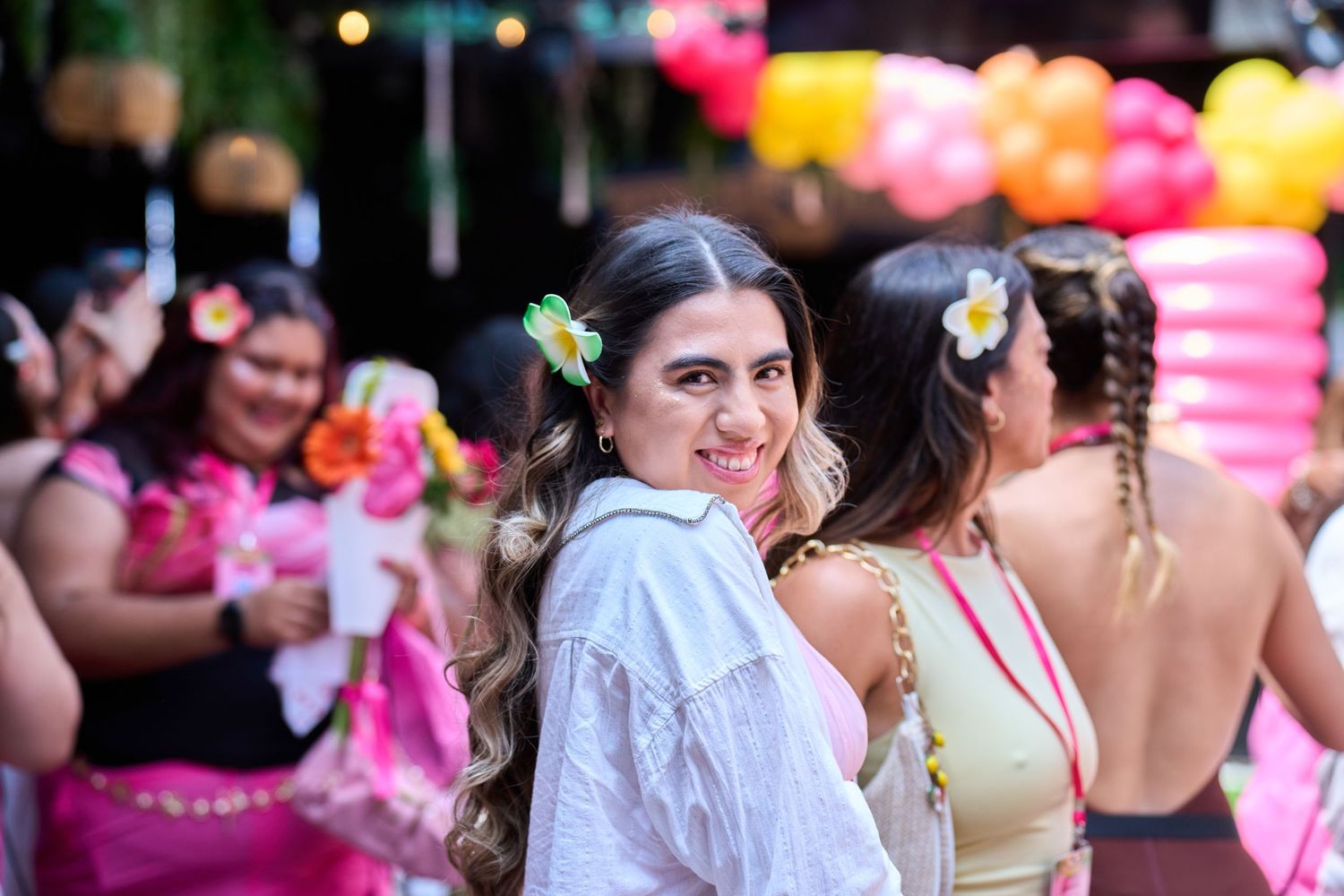 Woman with flower hair accessory smiling at tropical party with colorful balloons and guests in festive attire.