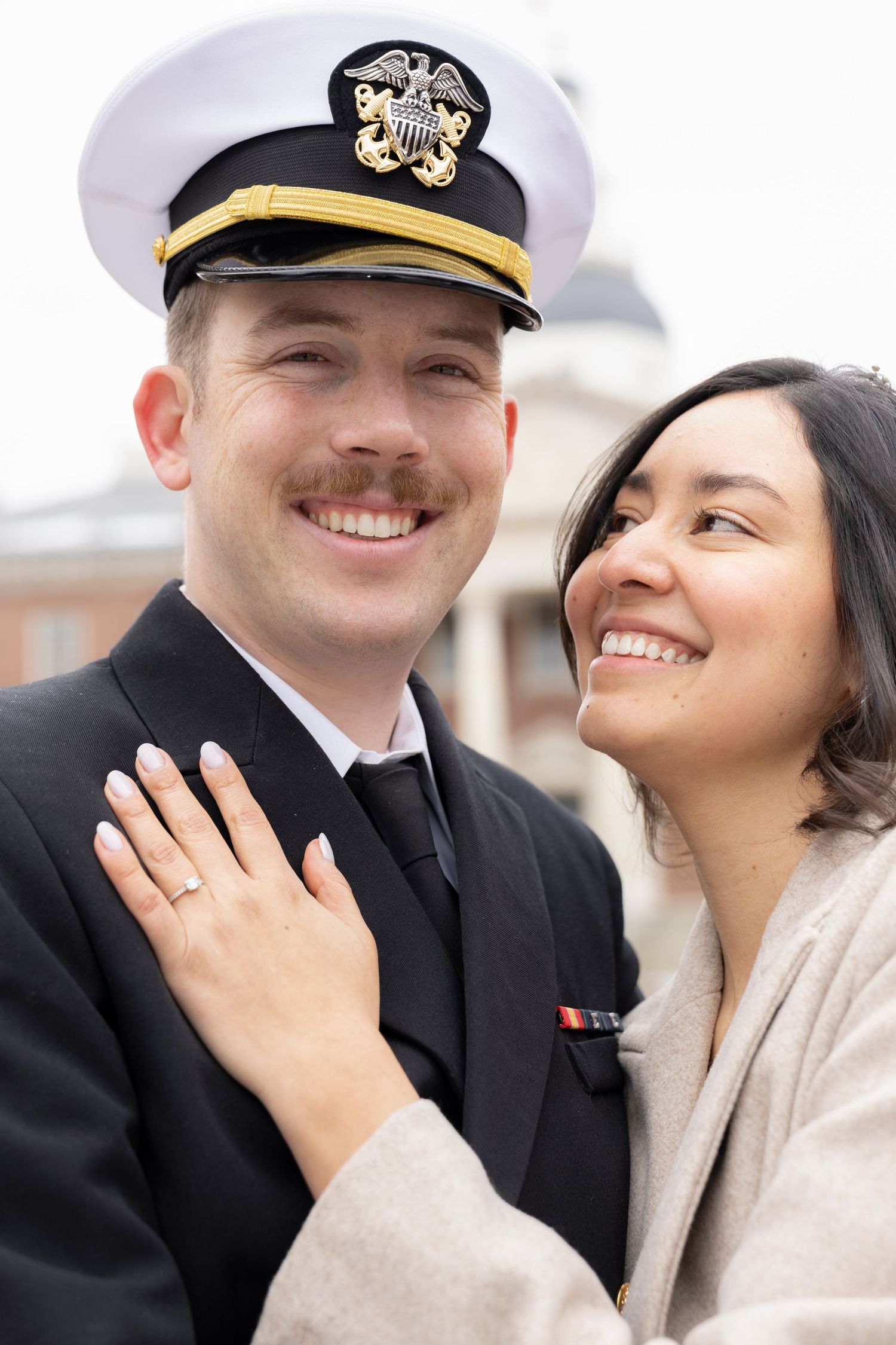 A happy engaged couple embraces outdoors, with the groom in naval dress uniform.