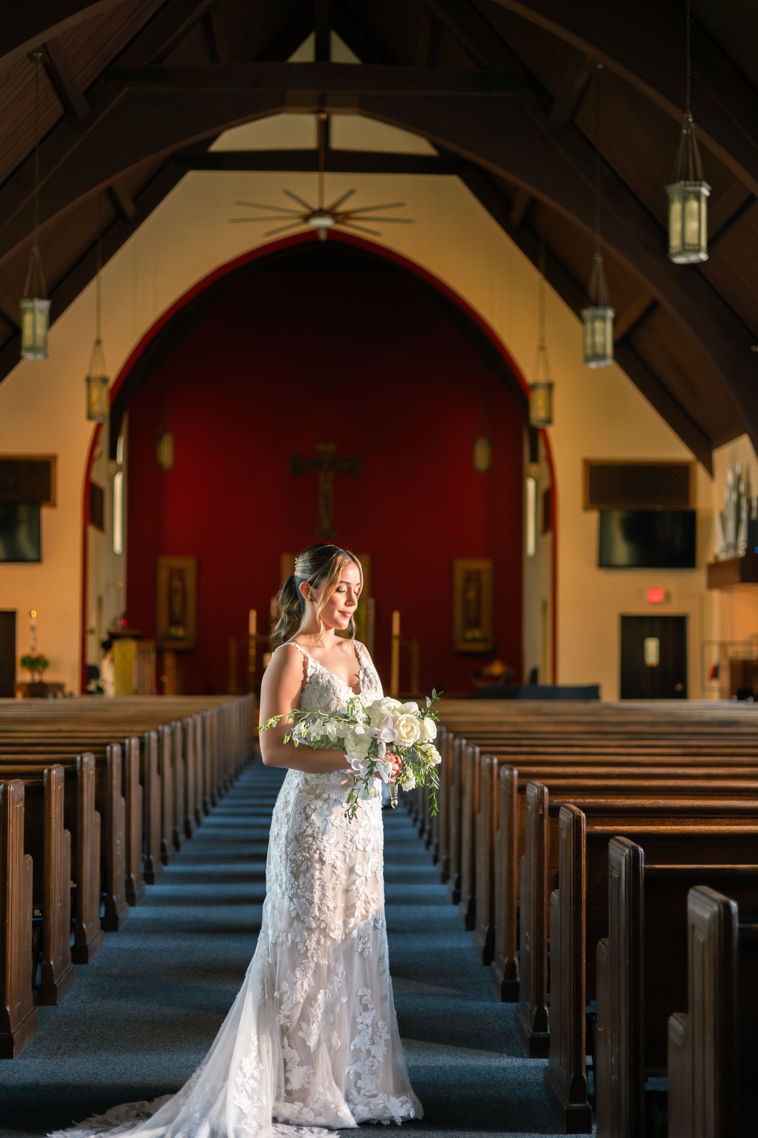 A bride in a flowing lace wedding dress walks gracefully through the aisle of a traditional church sanctuary.