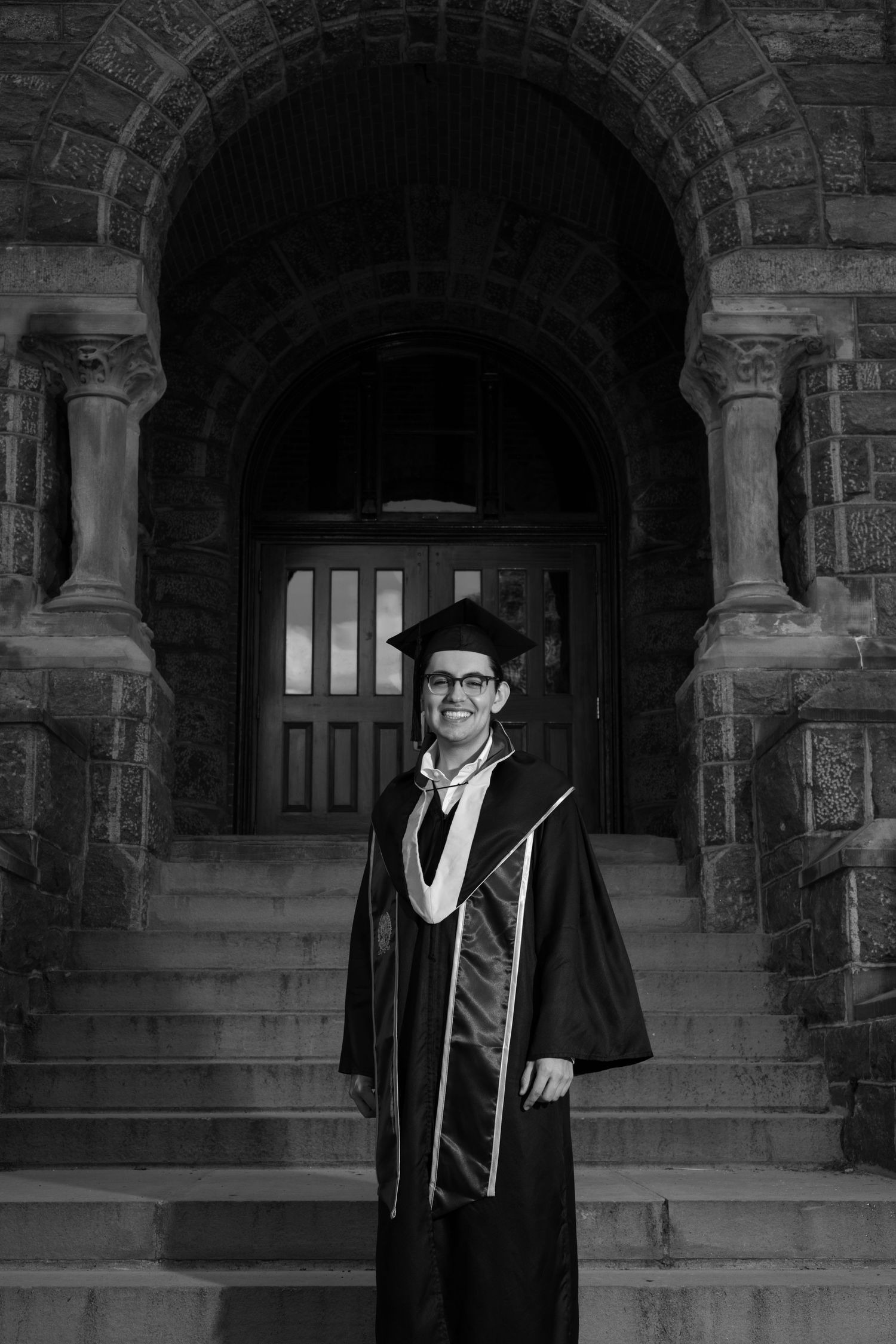 A graduate in traditional academic regalia stands on stone steps beneath an arched doorway in black and white.