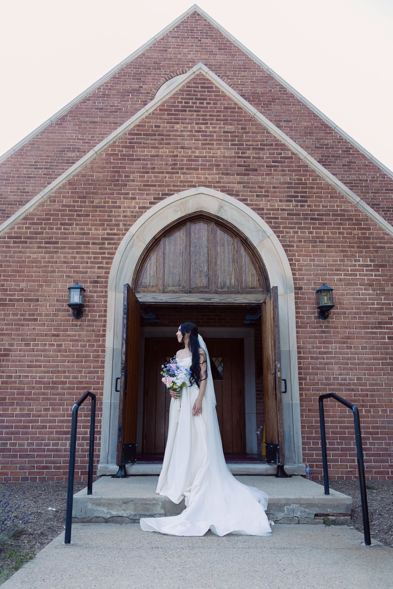A figure in a white wedding gown and long veil stands in the arched doorway of a brick church entrance.