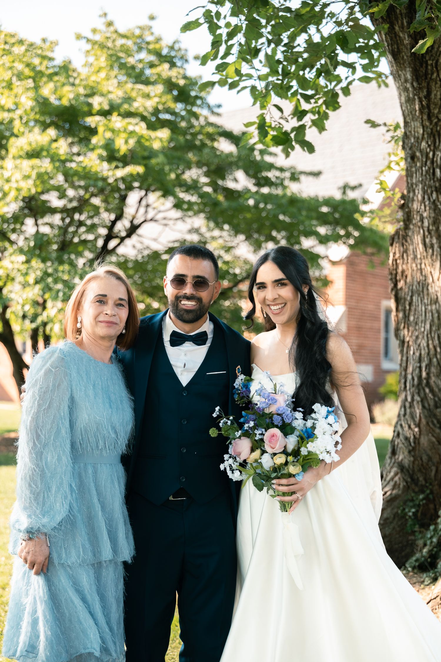 Three people pose together outdoors at a wedding with trees and greenery in the background.