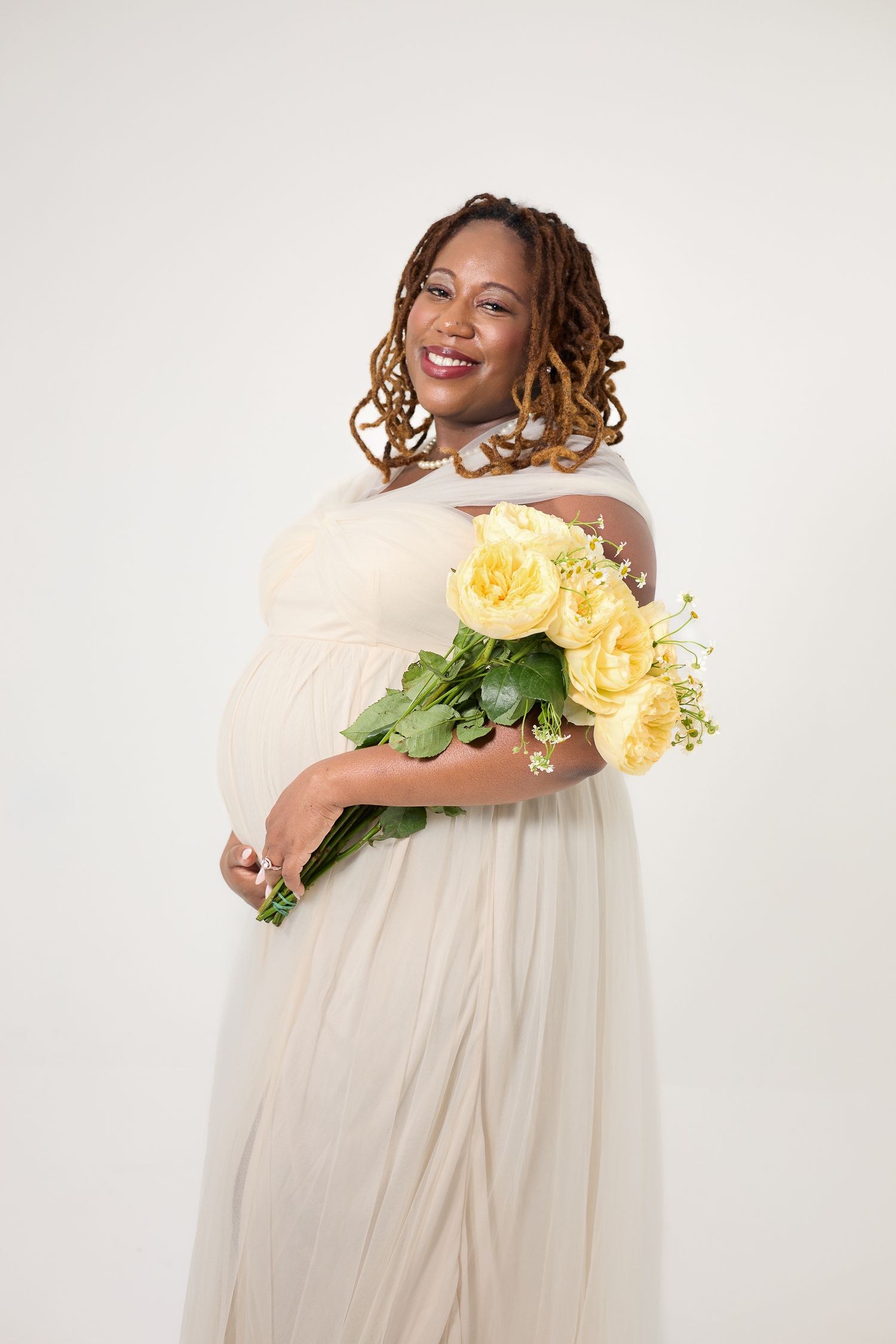Smiling pregnant woman in white dress holding yellow roses bouquet against white background.