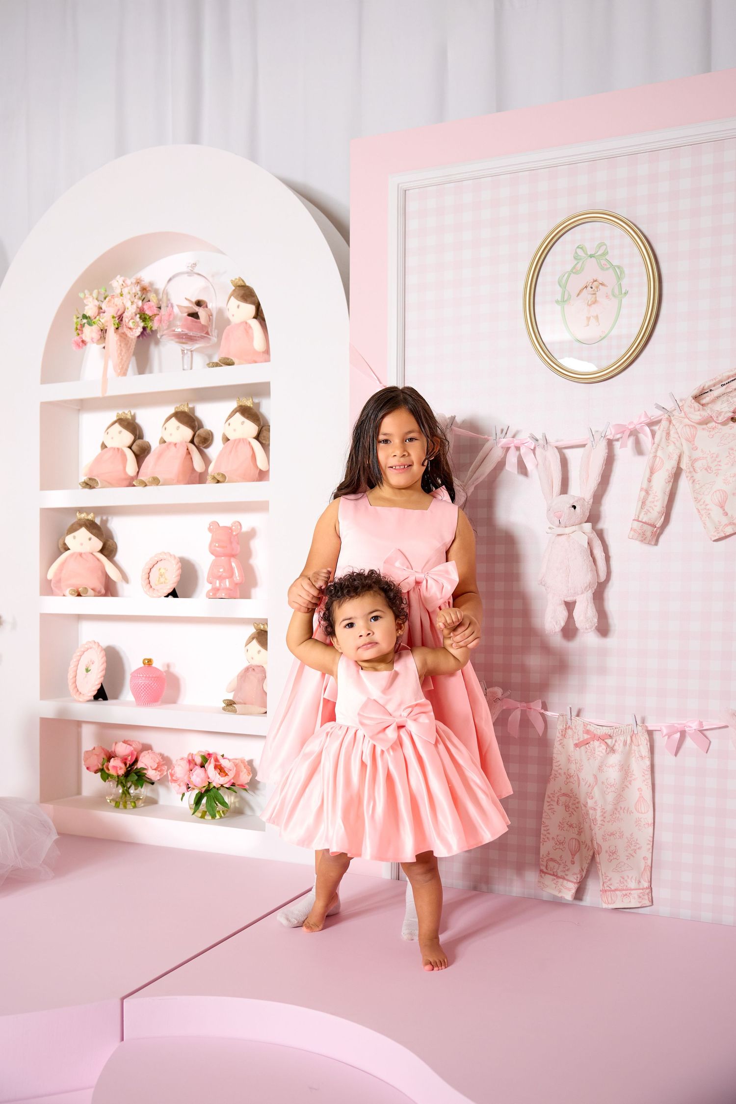 Two young girls in matching pink dresses pose in a pink decorated room with shelves of dolls and toys.