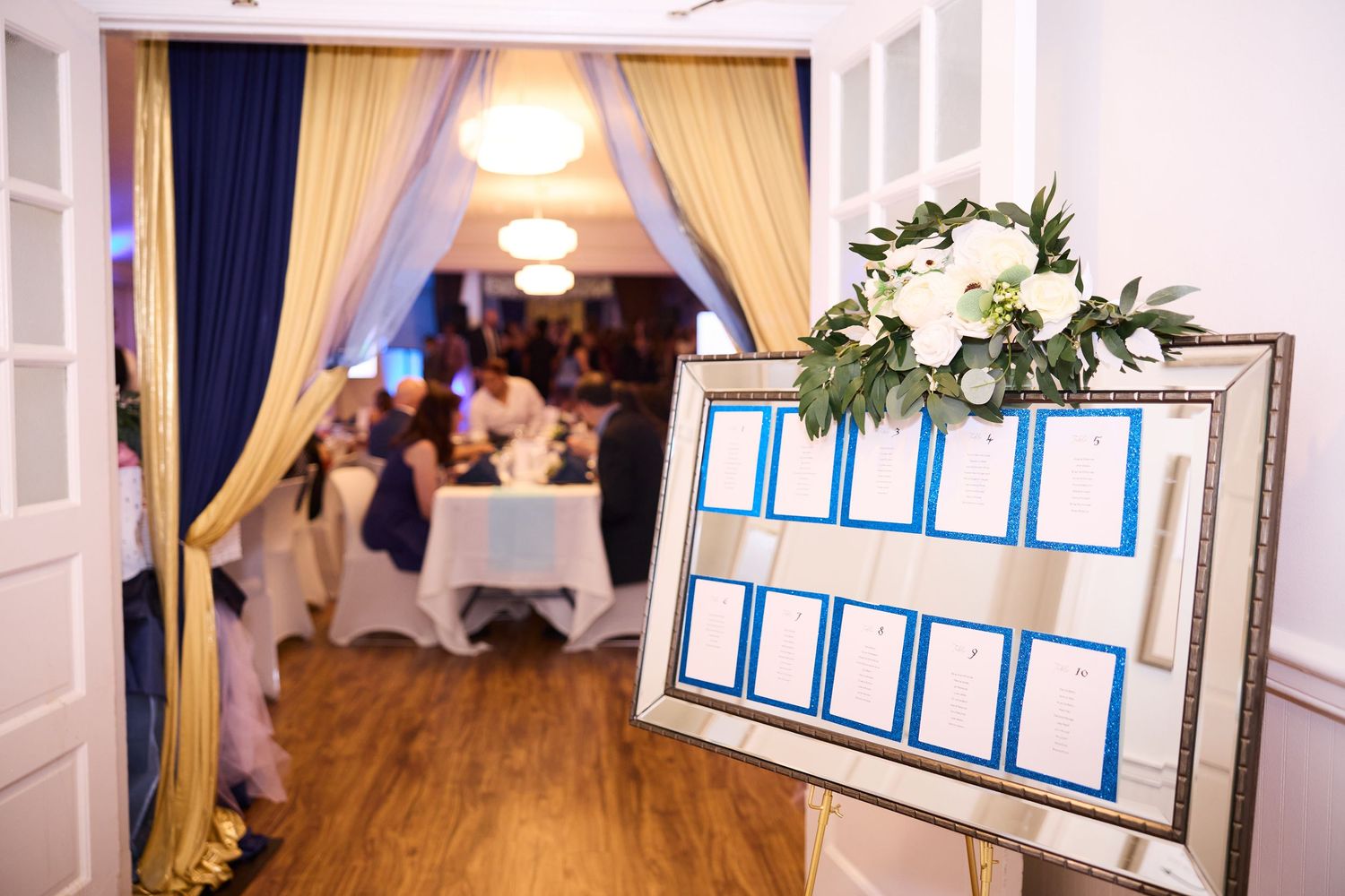 Elegant wedding venue interior with cream curtains, chandeliers and floral decorations along white walls.