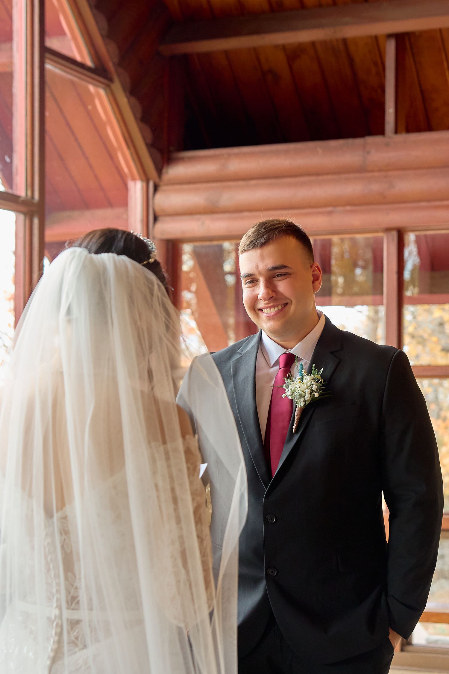 A wedding couple shares a moment in front of large windows in a rustic wooden chapel.