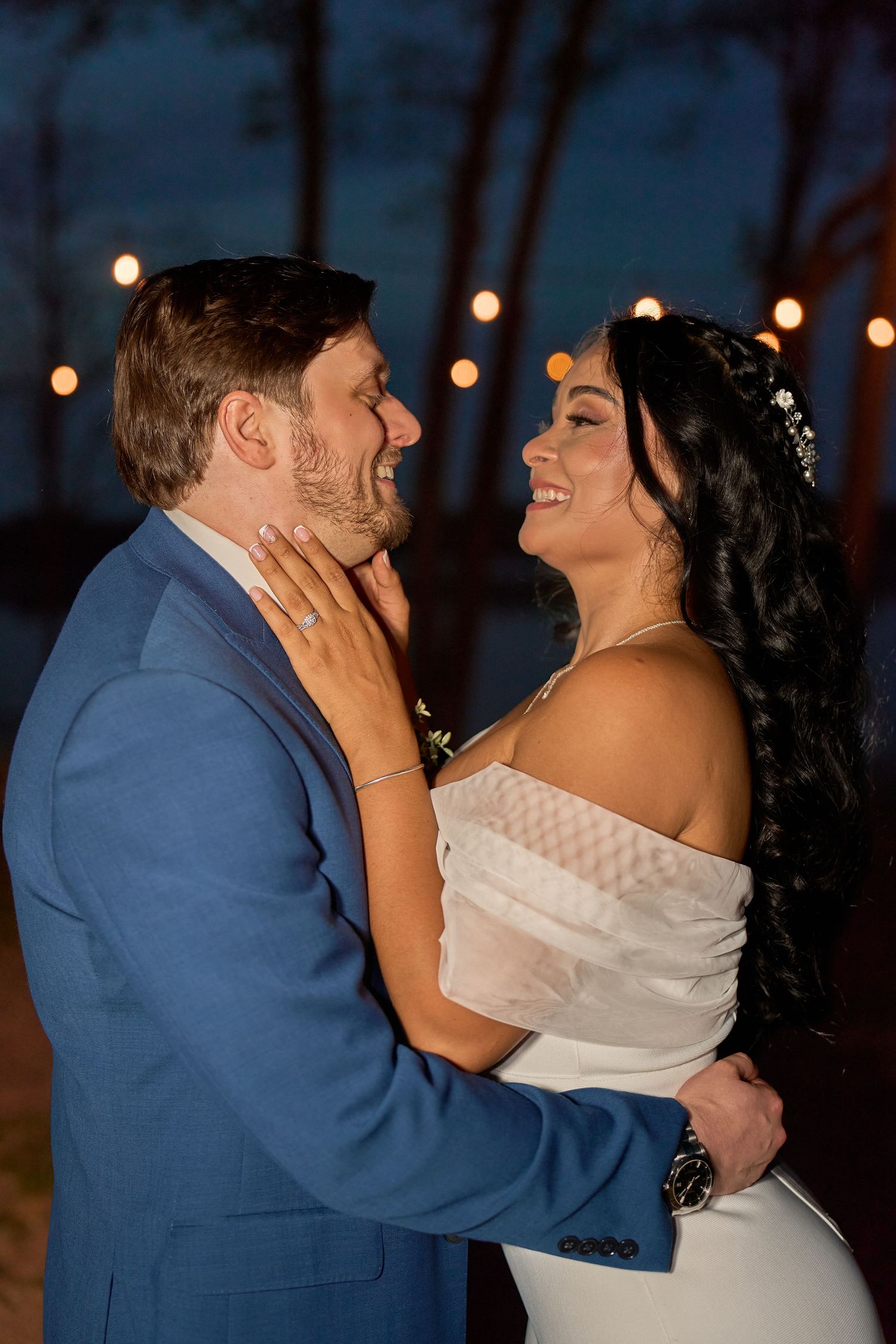 Bride and groom share a romantic moment at night, smiling at each other under warm string lights at their wedding.