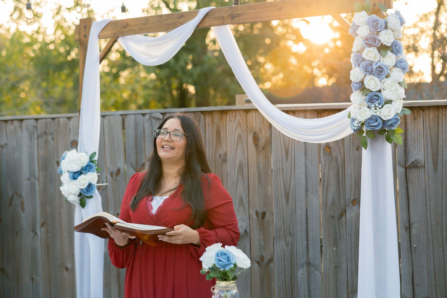 A person in a red sweater stands by a wooden fence decorated with white fabric and blue floral accents at an outdoor event.