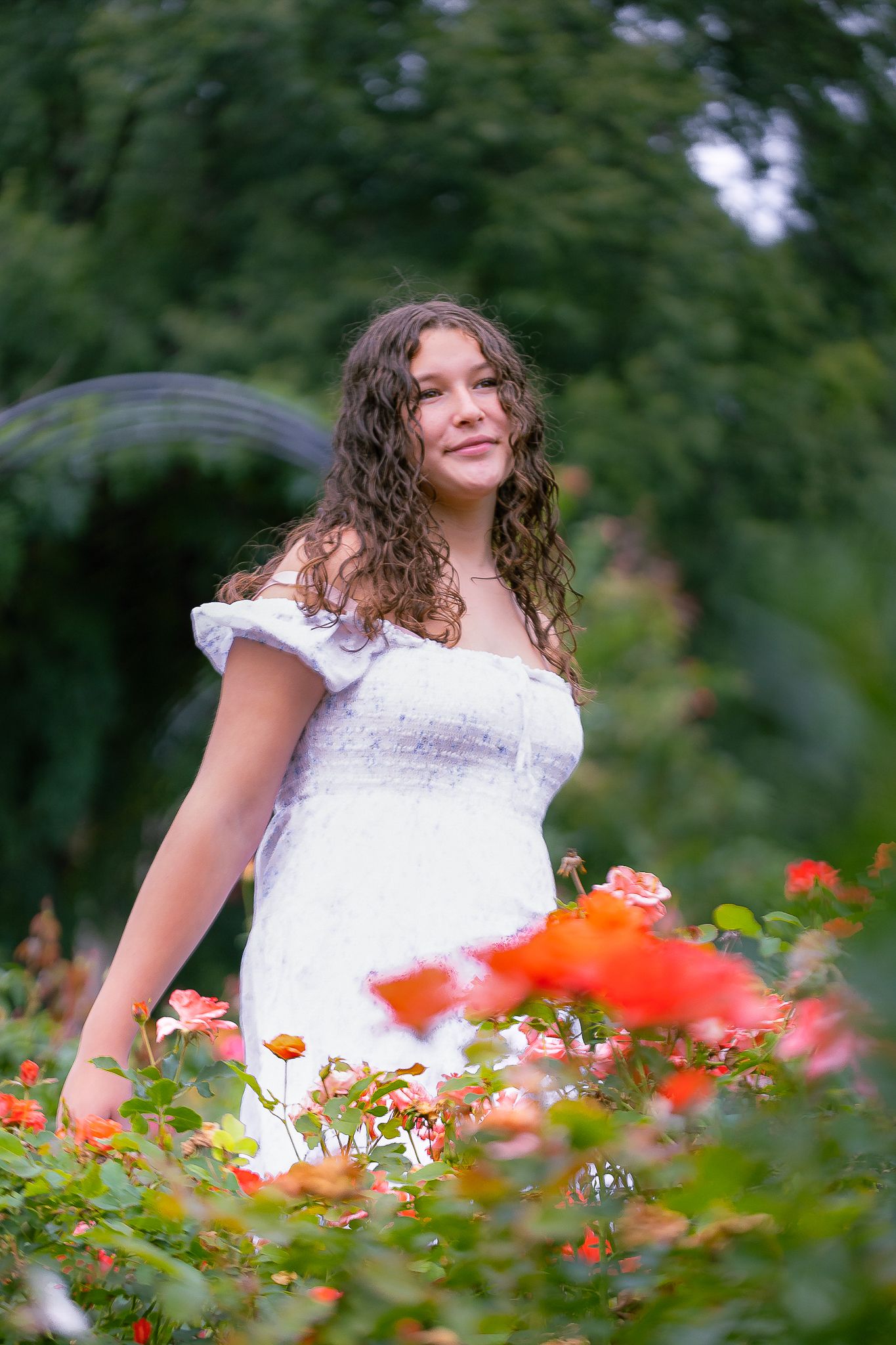 Person in white off-shoulder dress stands among blooming red flowers in a lush garden setting.