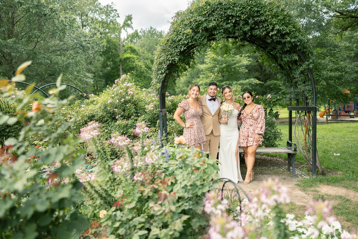 Wedding party poses beneath green garden archway surrounded by blooming flowers and foliage.