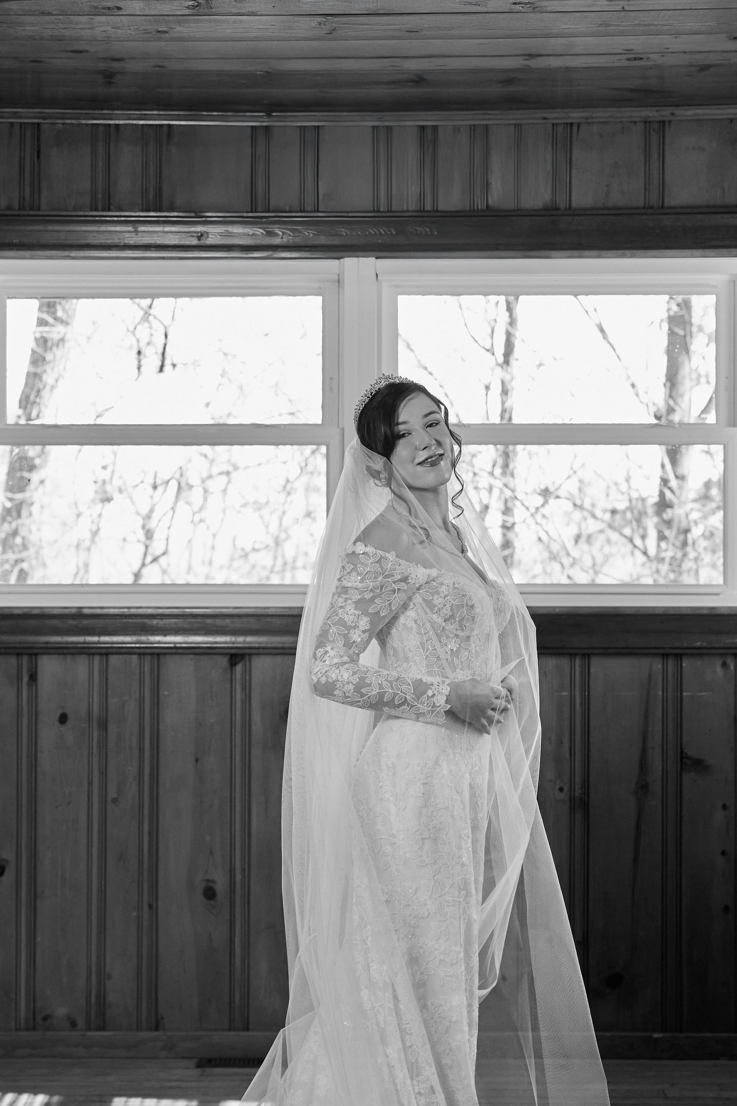 A black and white photo of a bride in a long sleeve lace wedding dress and veil standing by a window.