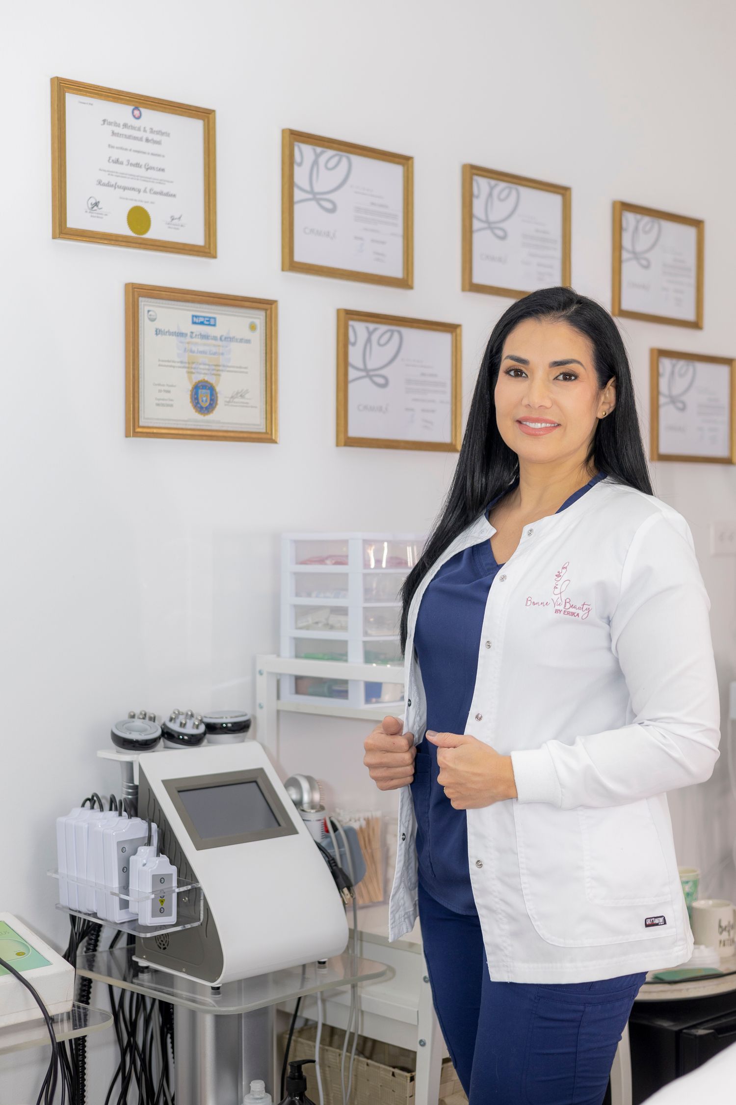 Healthcare professional in white lab coat stands in medical office with certificates and equipment visible in background.