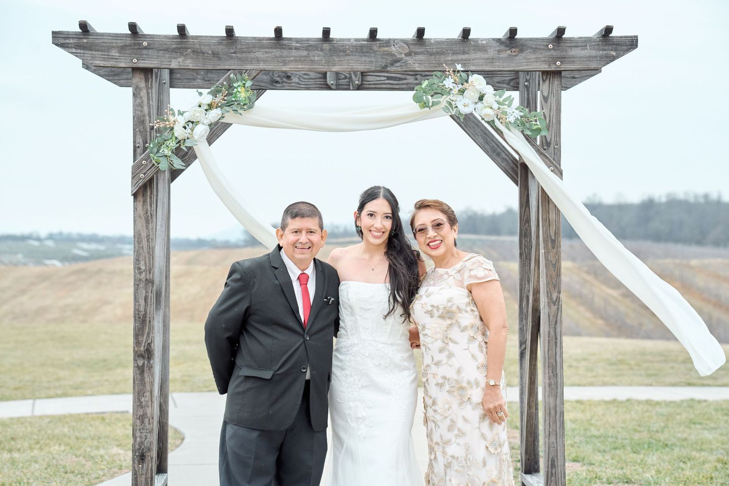 Wedding party gathered under a wooden arbor with scenic countryside views in the background.