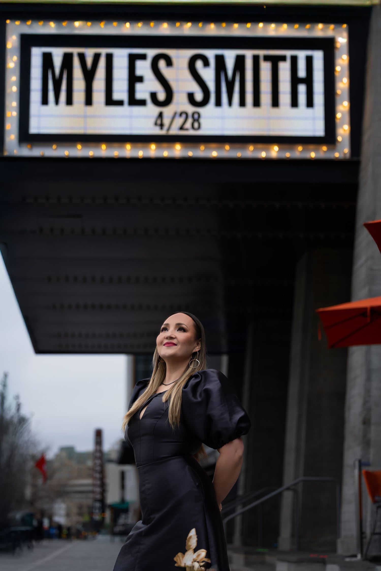 Woman poses beneath illuminated theater marquee sign at dusk.