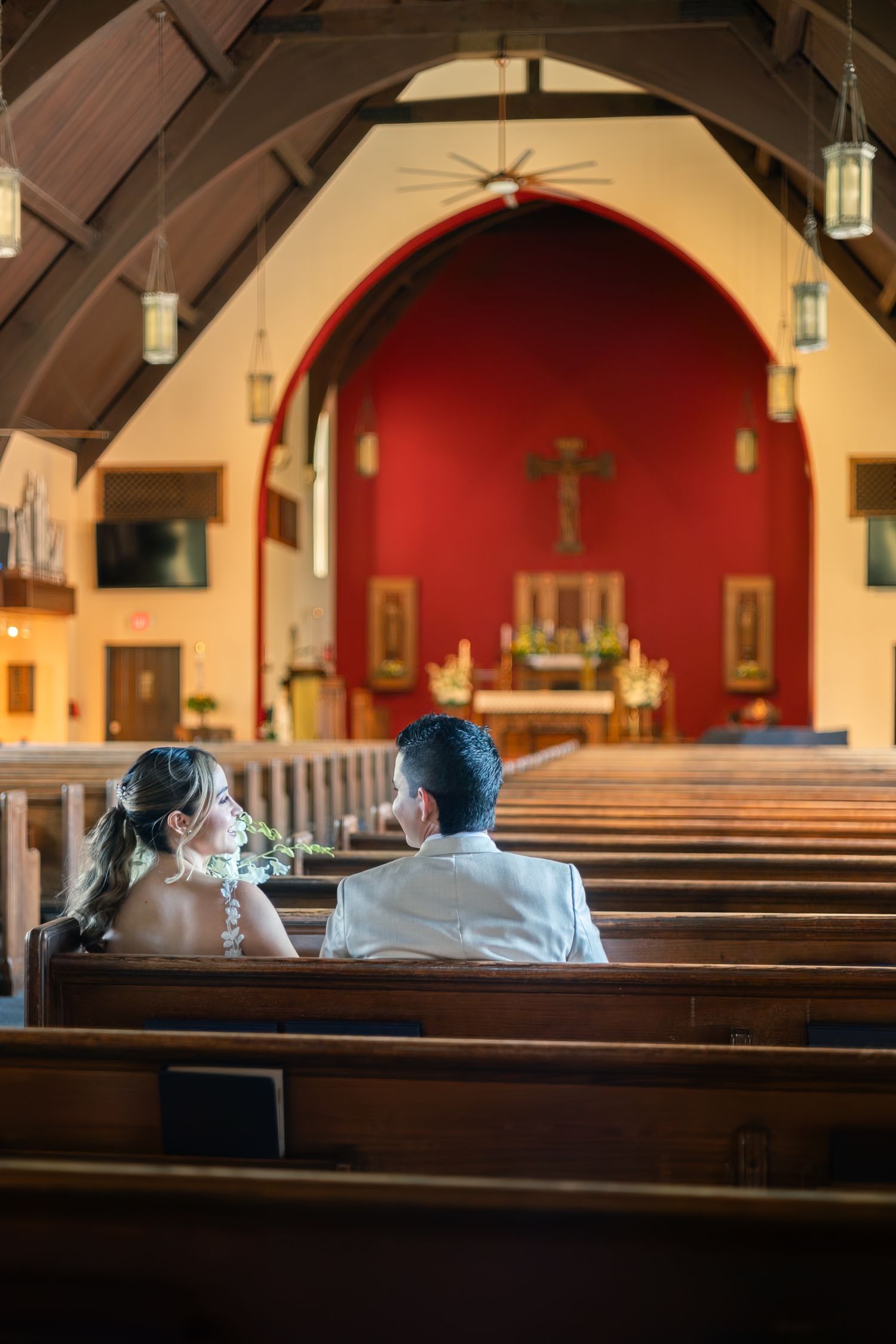 Two people sit in a church pew while light streams through stained glass windows in a traditional church interior.