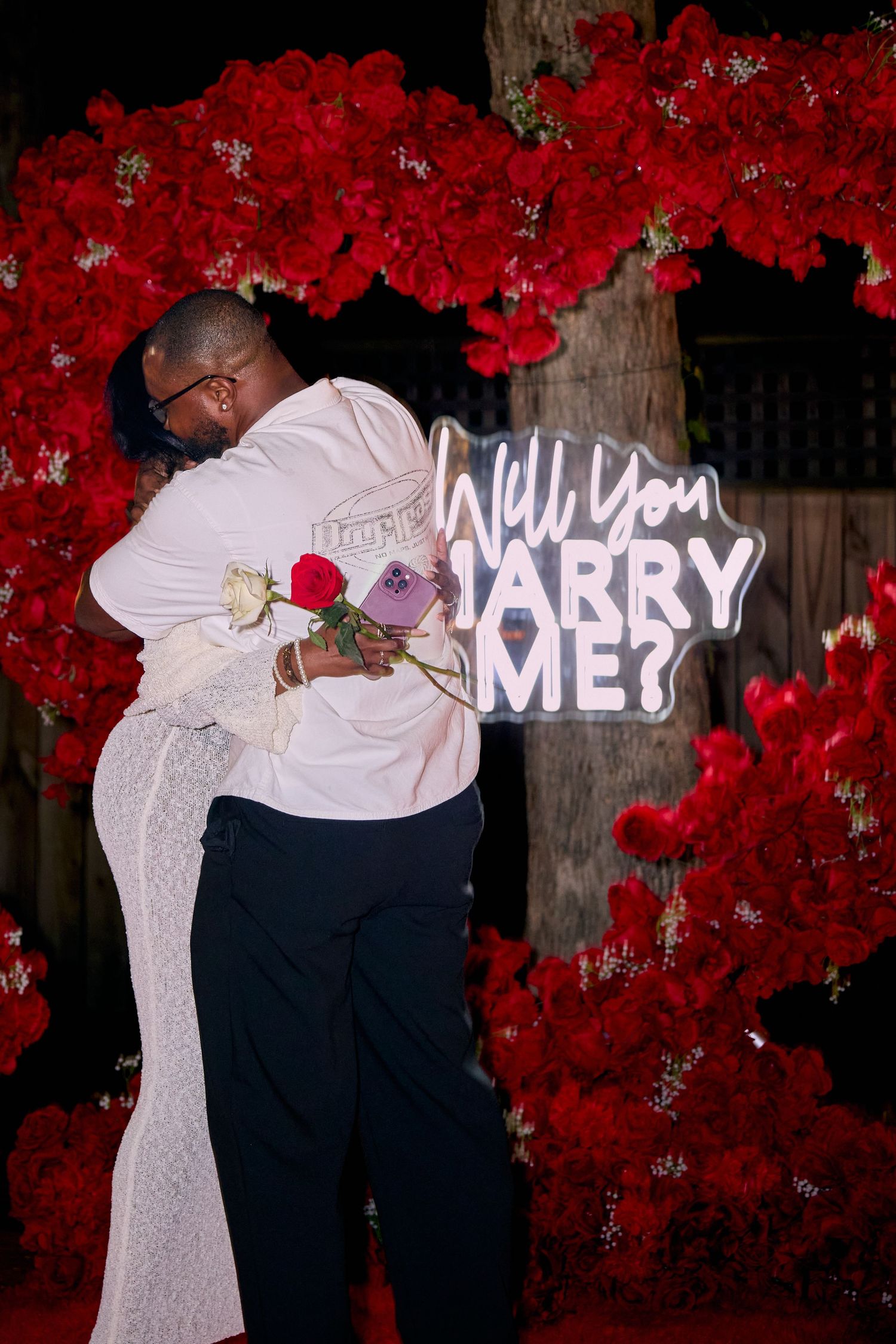 A romantic marriage proposal setup with a neon 'Will You Marry Me?' sign against a backdrop of vibrant red flowers.