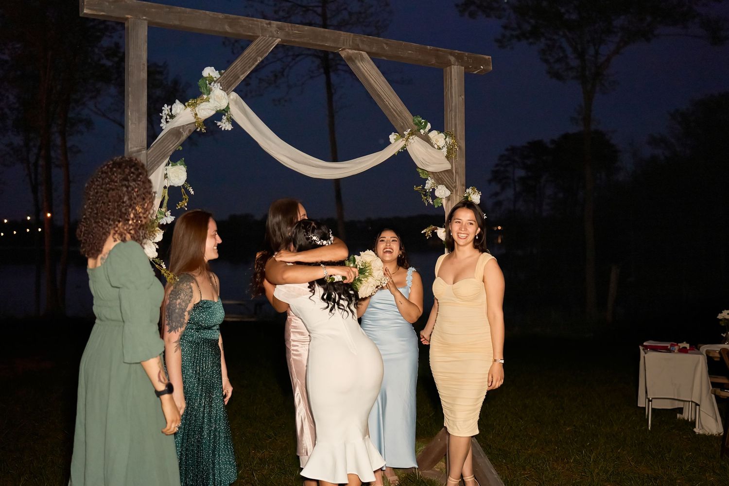 Women in elegant dresses celebrate at an outdoor wedding reception under a floral arch at night near a lake.