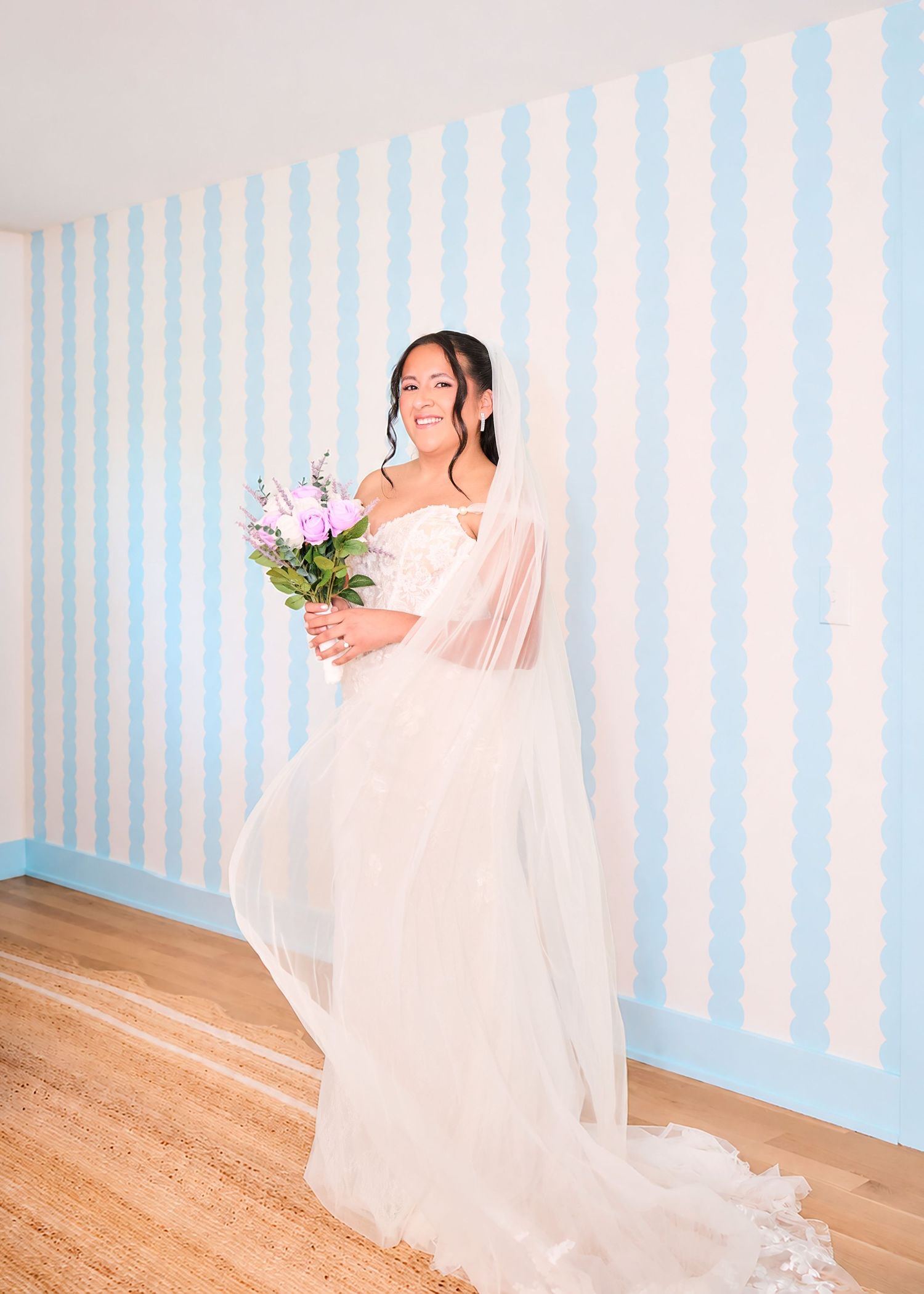 Bride in white gown holding purple bouquet poses in front of blue and white striped wall on hardwood floor.