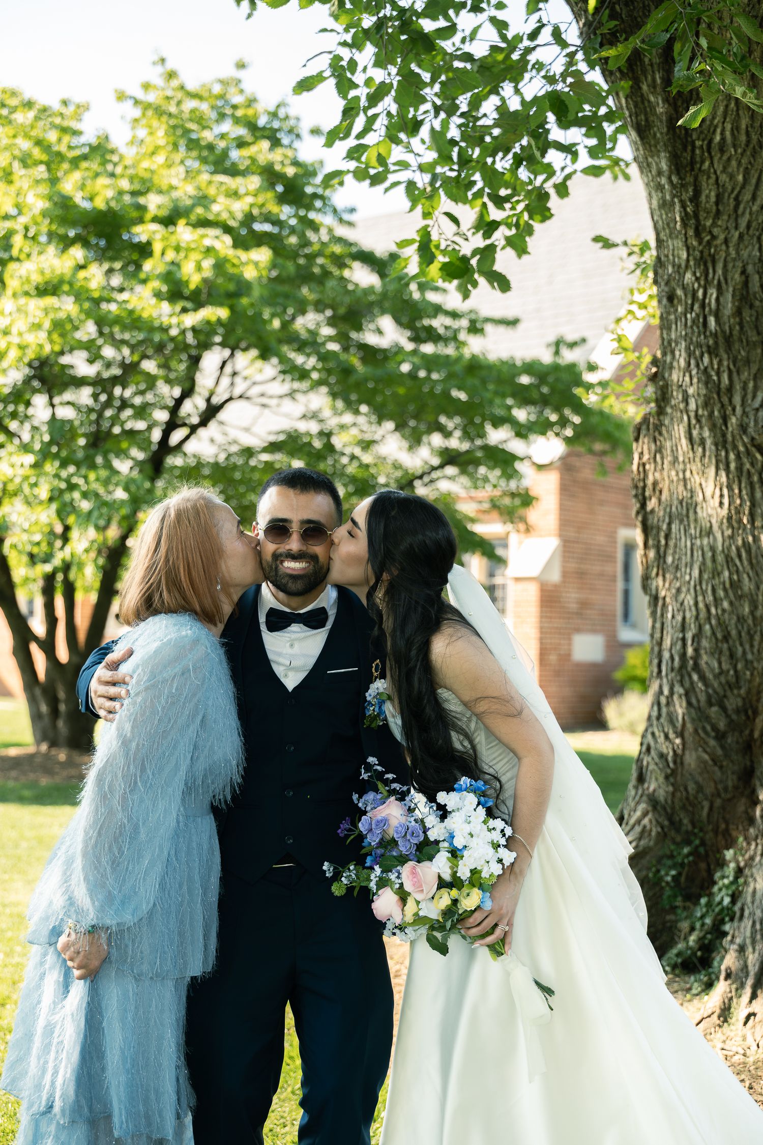 A couple embraces during an outdoor wedding ceremony under green trees with a bouquet of blue and white flowers.