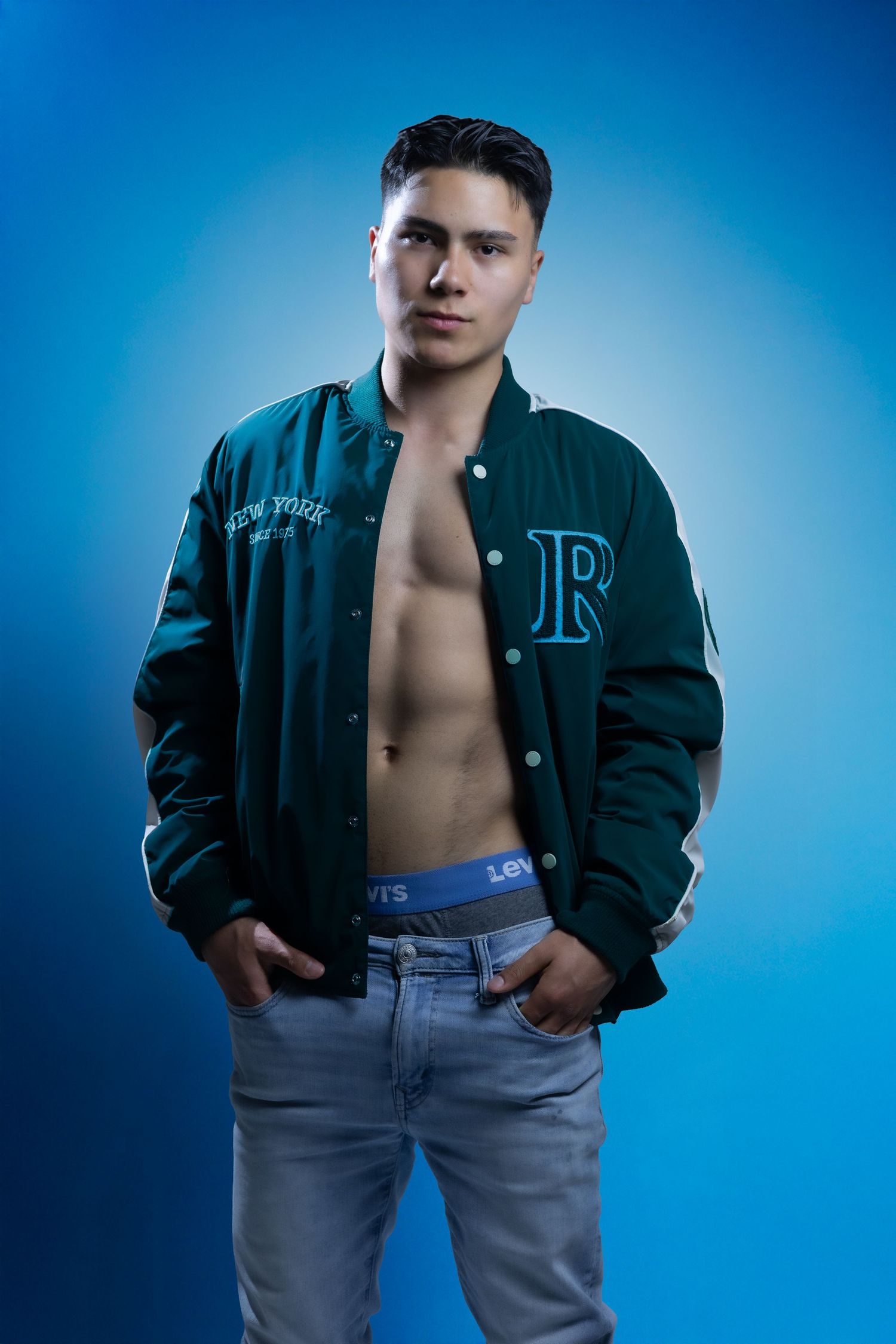 Athletic model poses against blue backdrop wearing an open varsity jacket and light denim jeans in studio lighting.