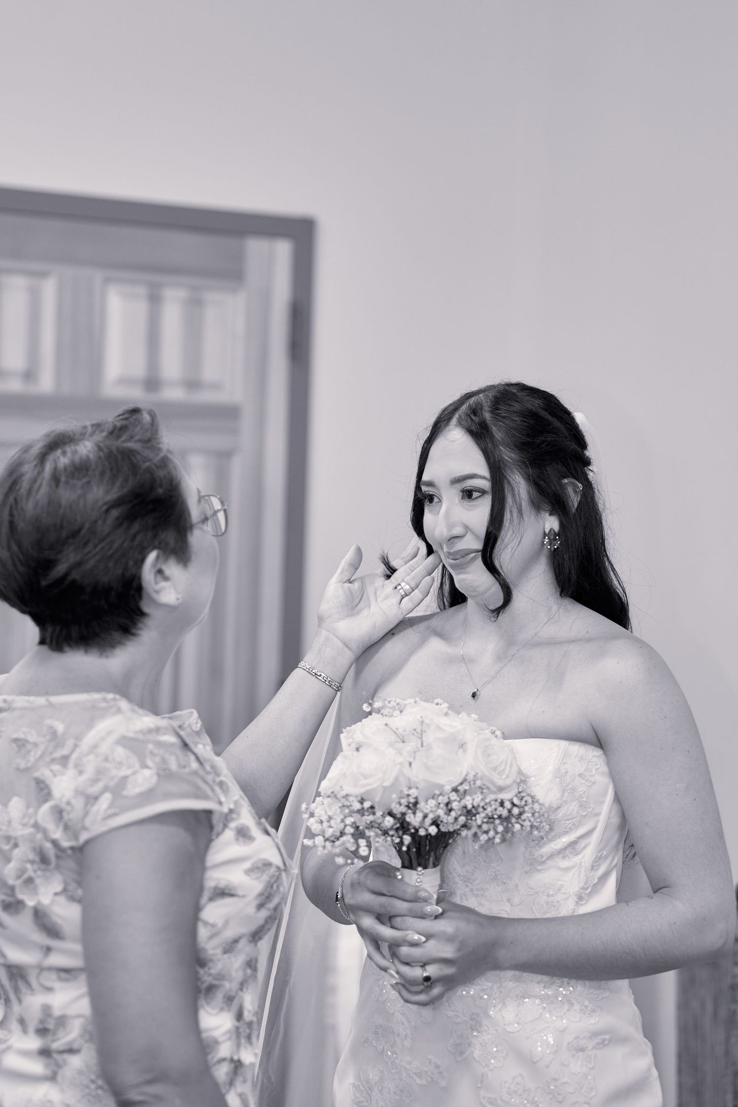 A bride in a white strapless gown holds a bouquet while having her makeup applied during wedding preparations.