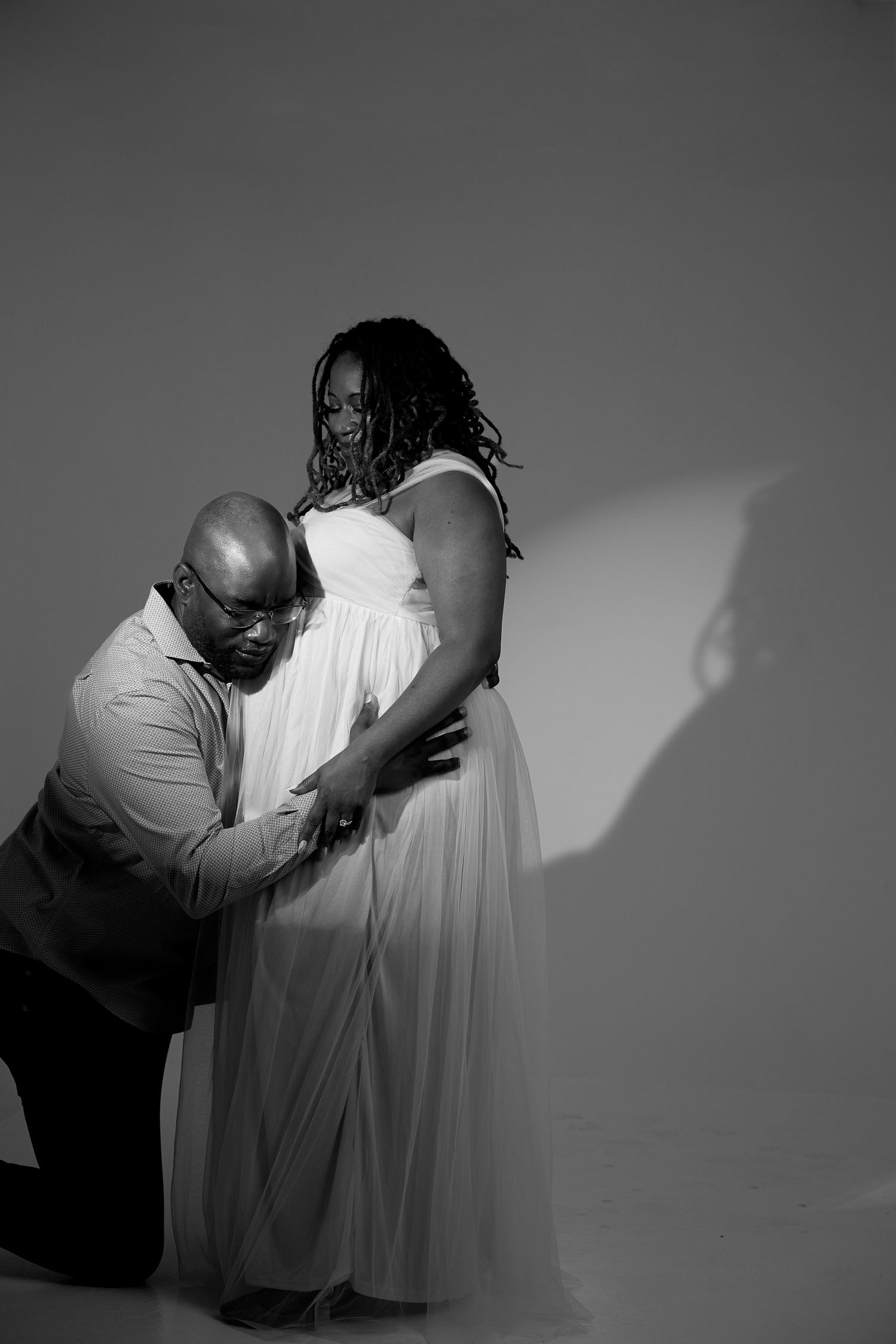 Black and white maternity photo of a couple, man kneeling kissing pregnant woman's belly in white gown.