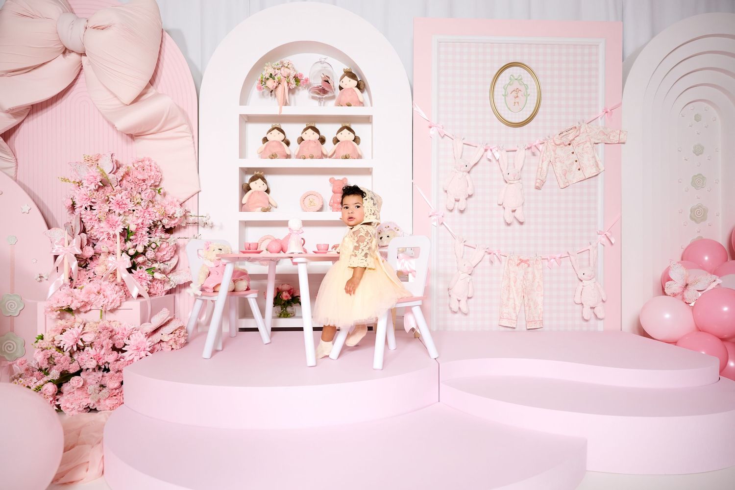 A young girl sits at a white table in a pink-themed party room with dolls, floral decor, and balloon accents.