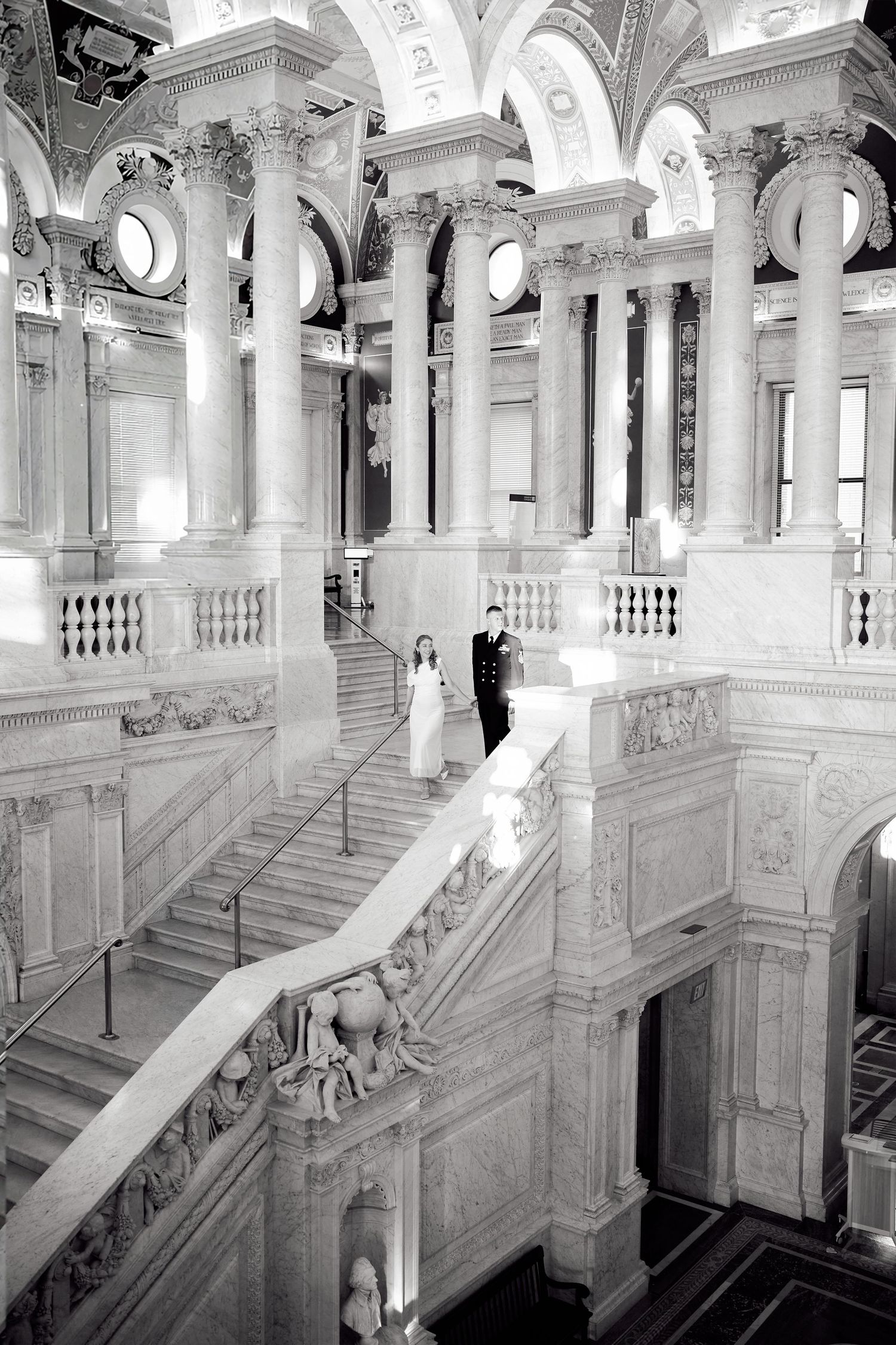 Black and white photo of bride and groom descending grand marble staircase in ornate neoclassical building.