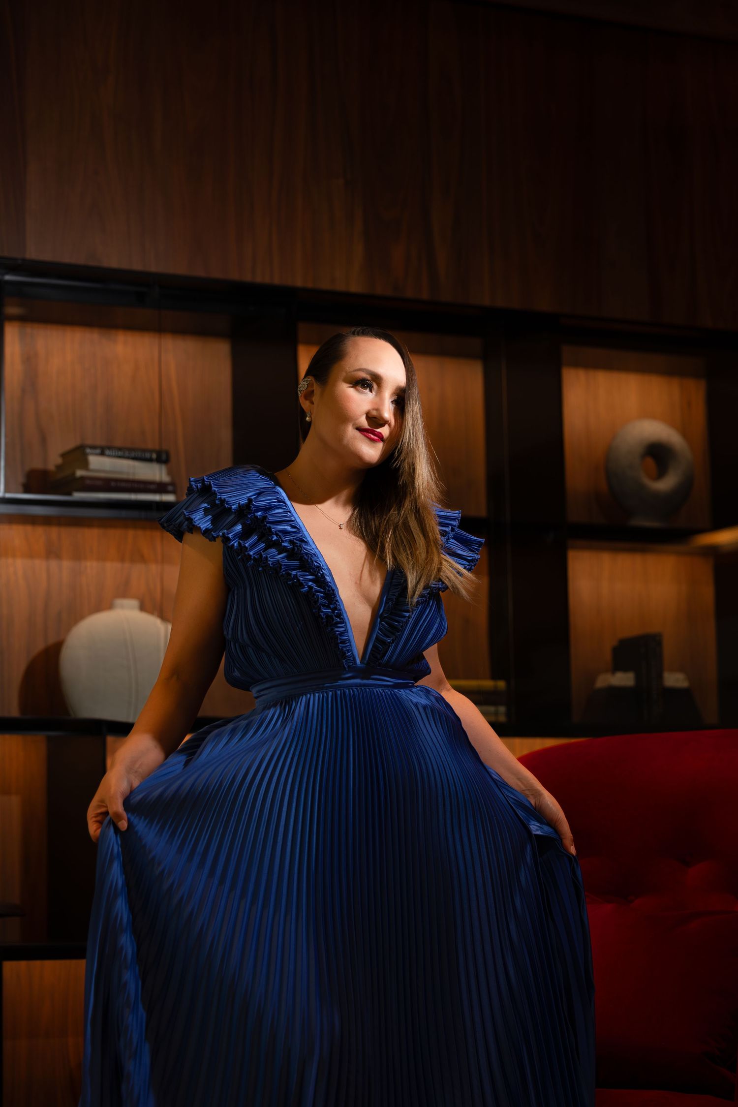 Pleated royal blue evening gown photographed in warmly lit interior setting.