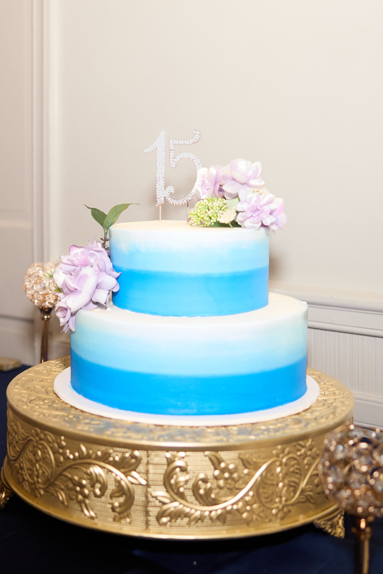 Two-tiered blue and white wedding cake with delicate floral decorations displayed on an ornate gold stand.