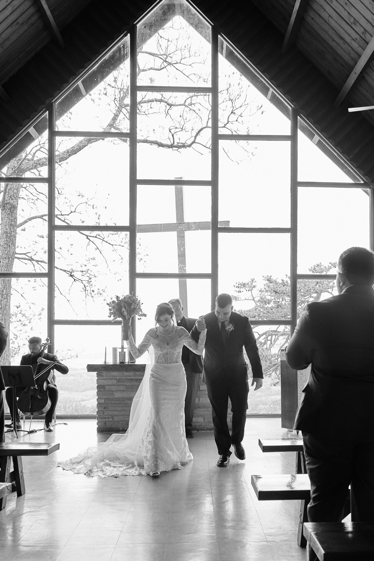 A couple walks down the aisle in a modern chapel with a dramatic triangular window and natural light streaming in.